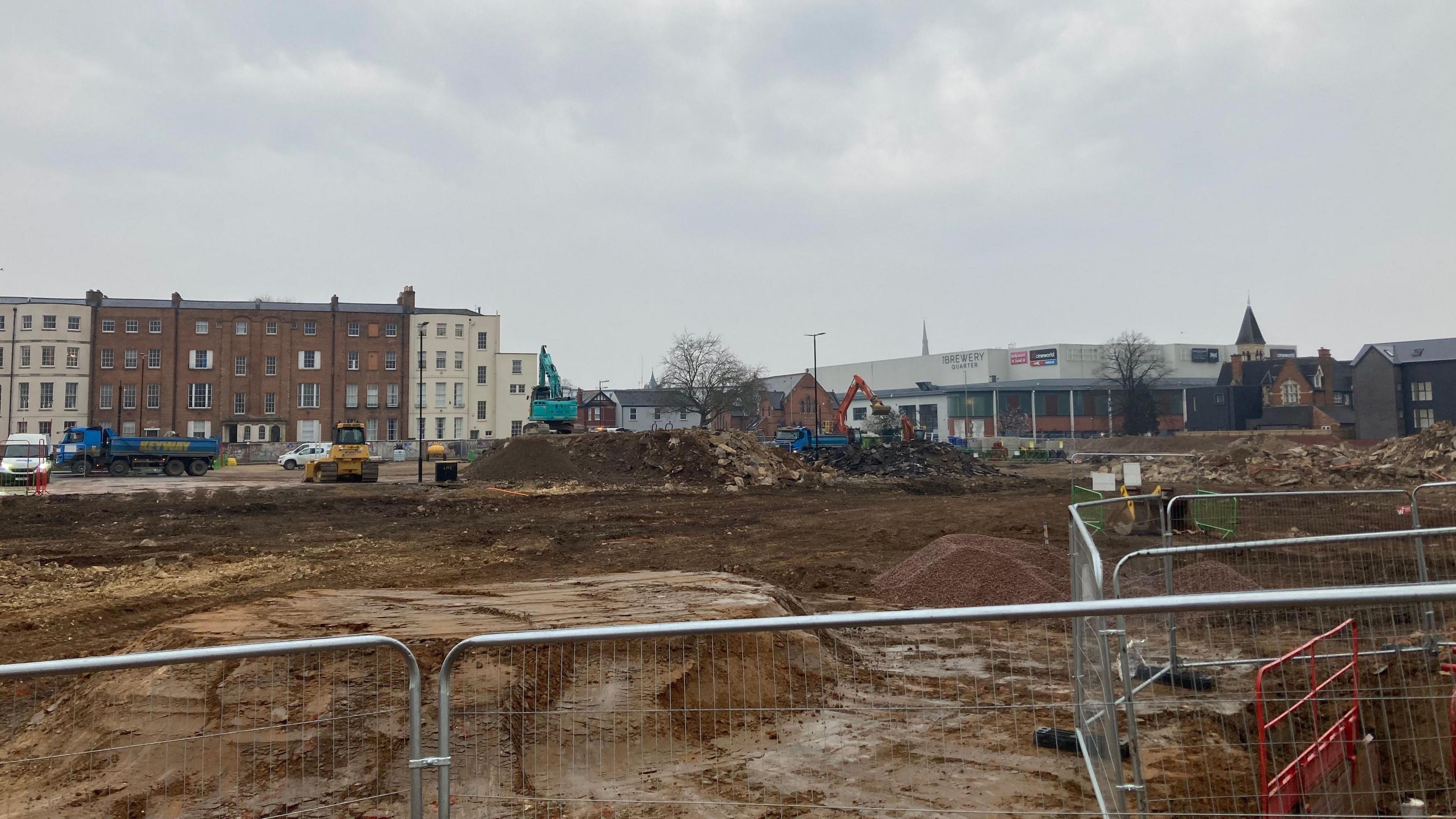 A brownfield site with mounds of dirt and rubble. Apartments and an industrial building can be seen in the background, with a road with several vehicles. There are metal fences in the foreground.