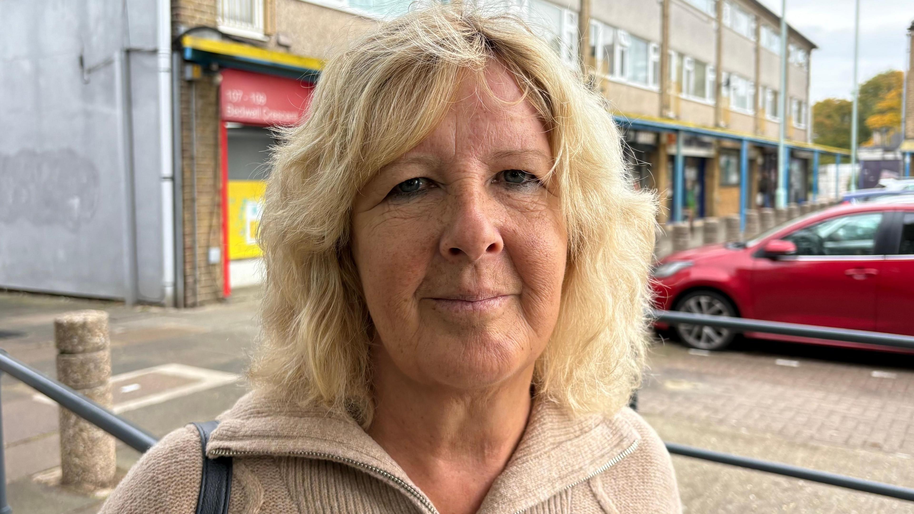 Deborah Johnston with long blond hair looking at the camera and wearing a light brown zip-up top. There is a parade of shops behind her with a car park to the right.
