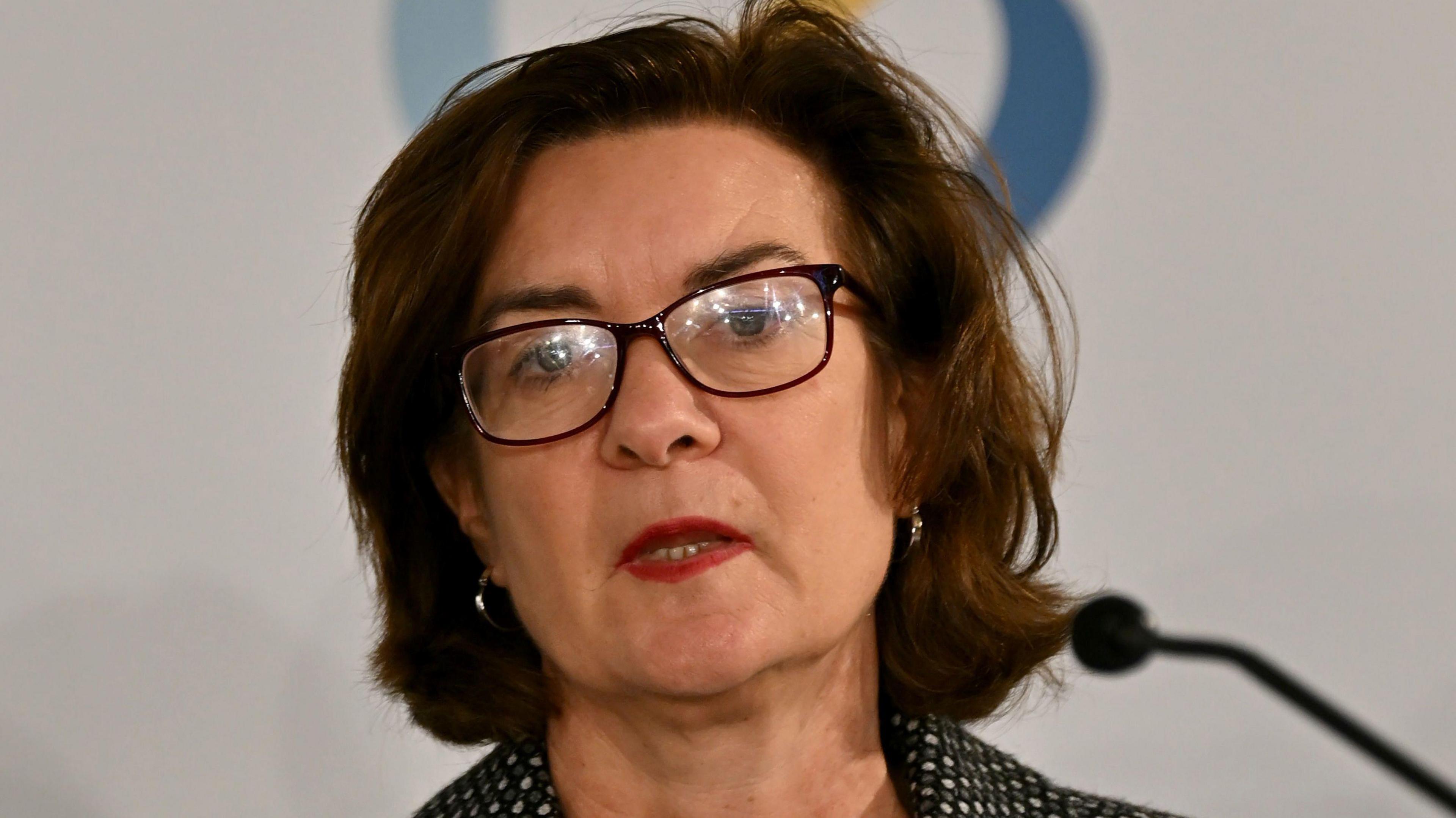 A head and shoulders picture of Eluned Morgan speaking into a microphone. She is wearing glasses, and a pokadot jacket.