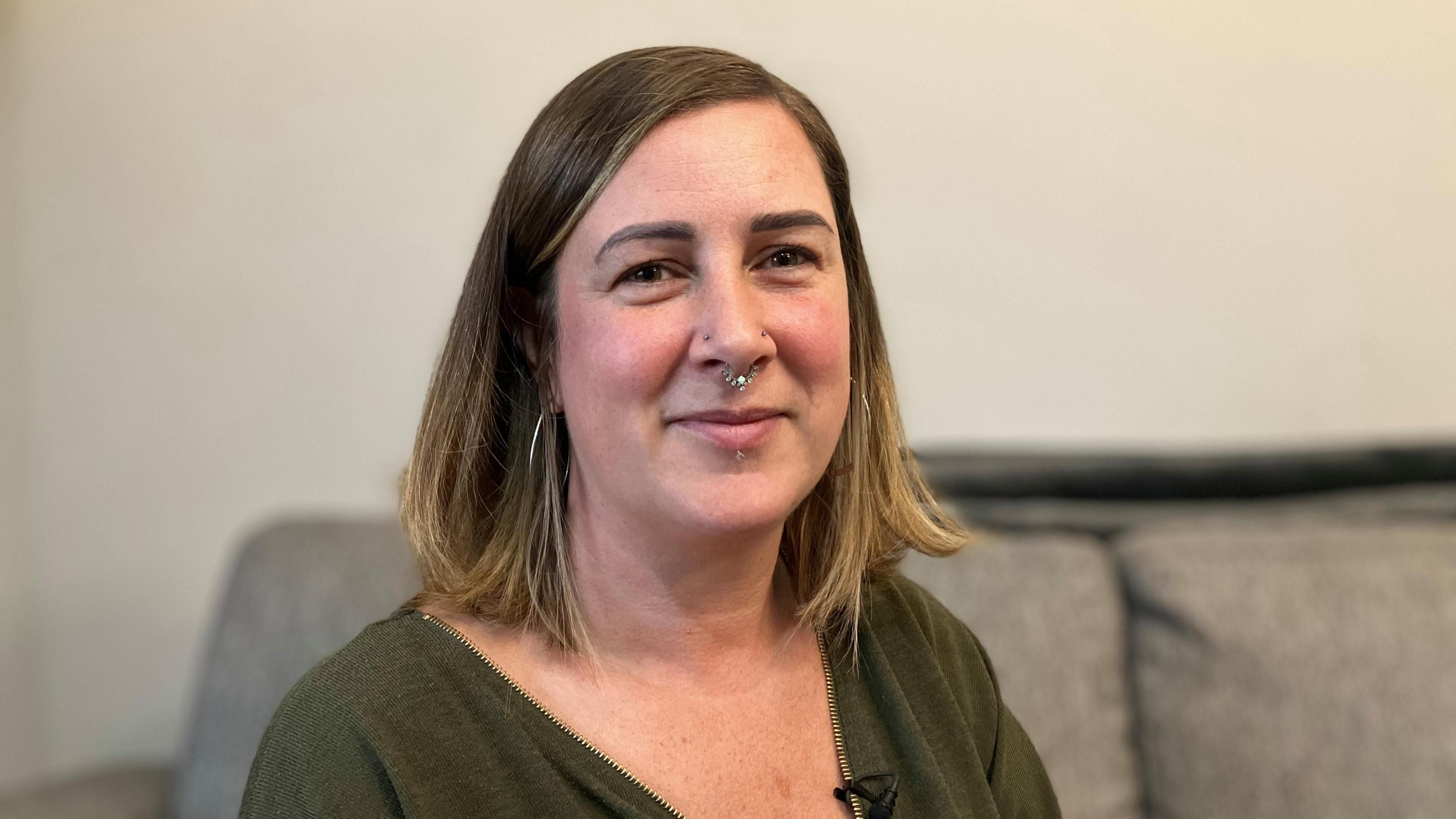 Gemma Barrett wearing a green top. She has a nose ring in and shoulder-length brown hair. She appears to be sitting in a home, with the back of a grey sofa visible in the blurred background.