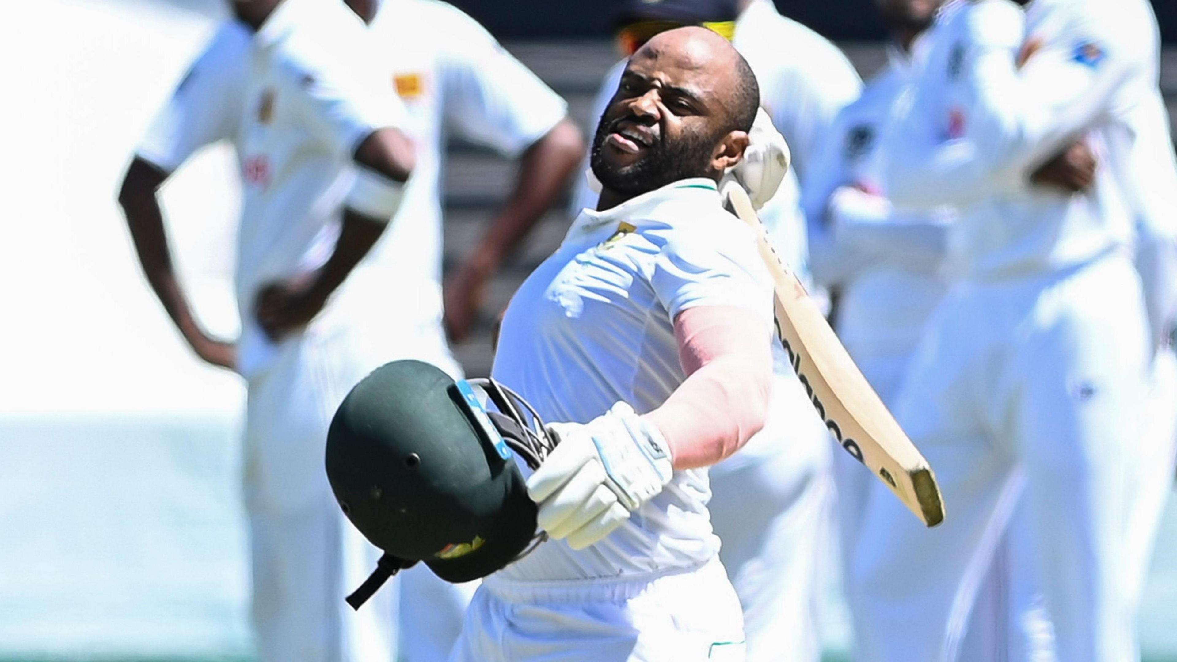 Temba Bavuma, wearing Test whites, holds his green helmet in his left hand and his cricket bat behind his head while celebrating after scoring a century against Sri Lanka in November 2024, while an out-of-focus group of fielders watch on in the background