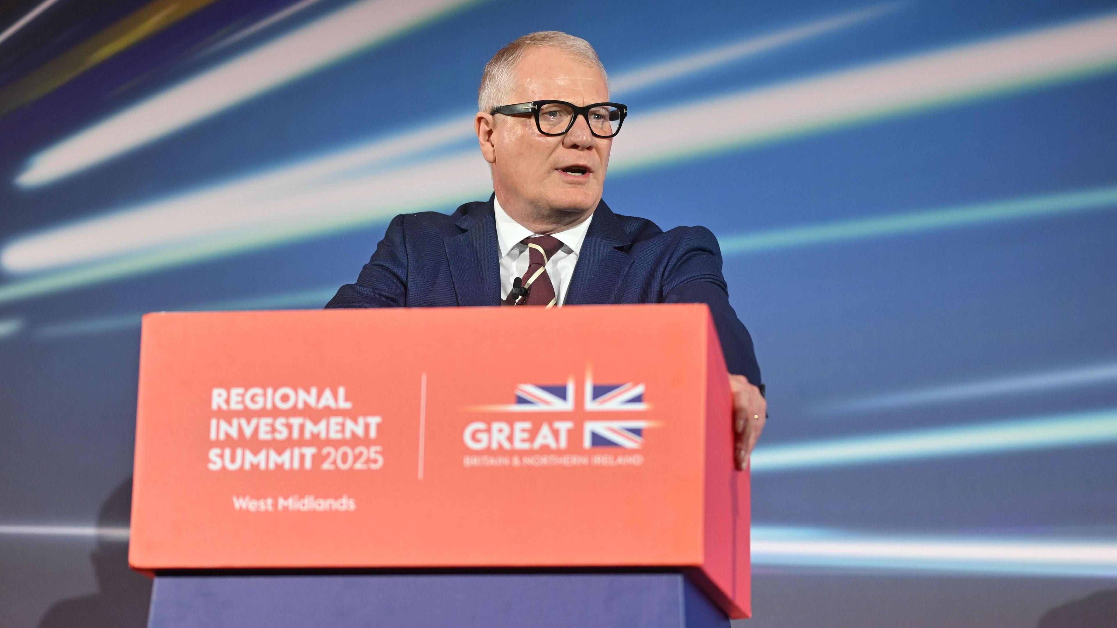 A man with short grey hair wearing a suit and thick black glasses, stood behind a red lectern with a red, white and blue Union Jack flag on it reading "Regional Investment Summit 2025 West Midlands".