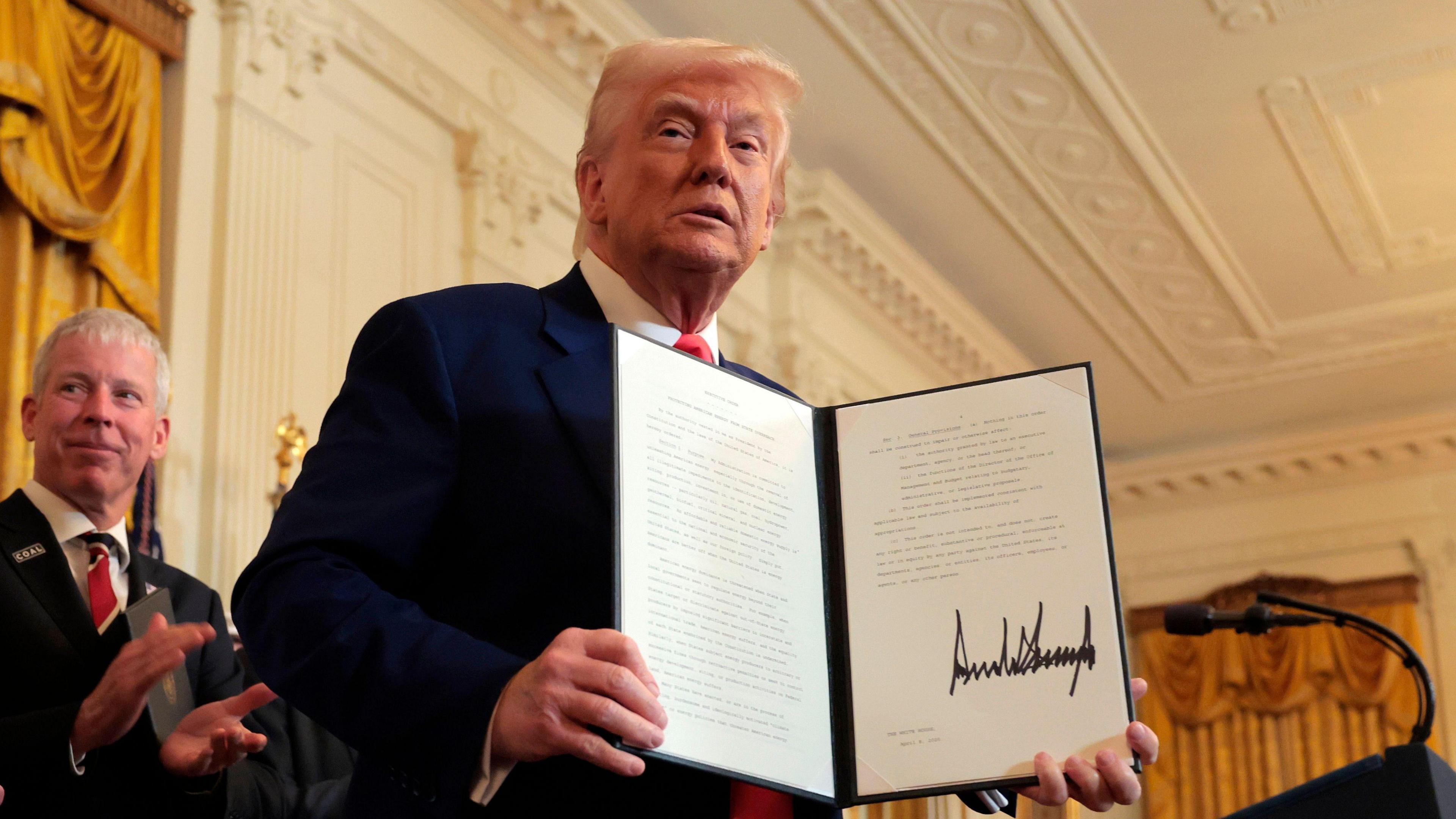 U.S. President Donald Trump holds up a signed executive order during a ceremony, with officials applauding in the background.