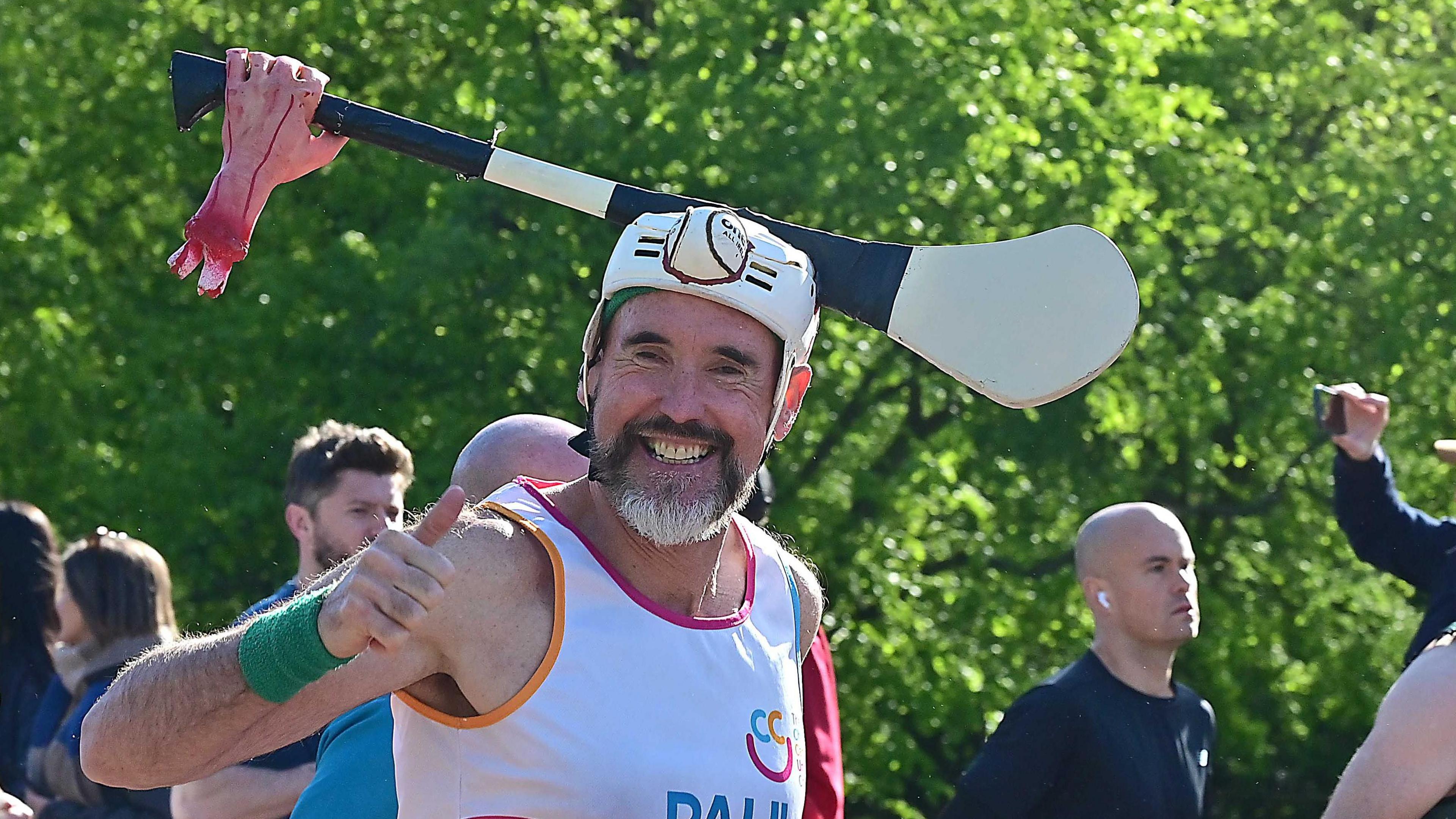A man with a black and grey beard smiling with his thumbs up as he runs past. He is wearing a helmet with a hurling bat stuck in it. He has a white running vest on and a green wristband. 