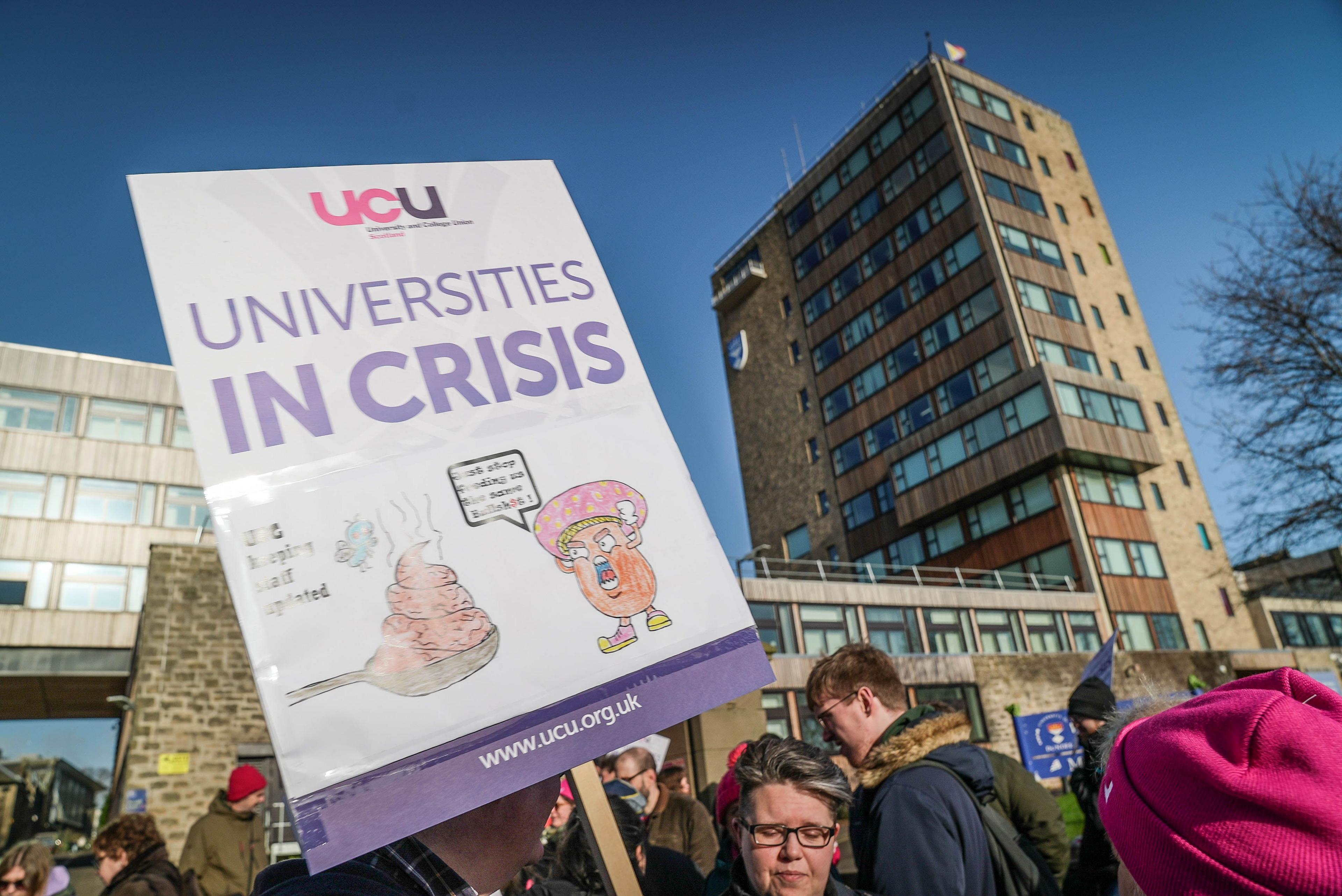 A protester holds a placard reading “Universities in Crisis” with the UCU logo during a demonstration outside a university building. Other people are gathered around under a clear blue sky.
