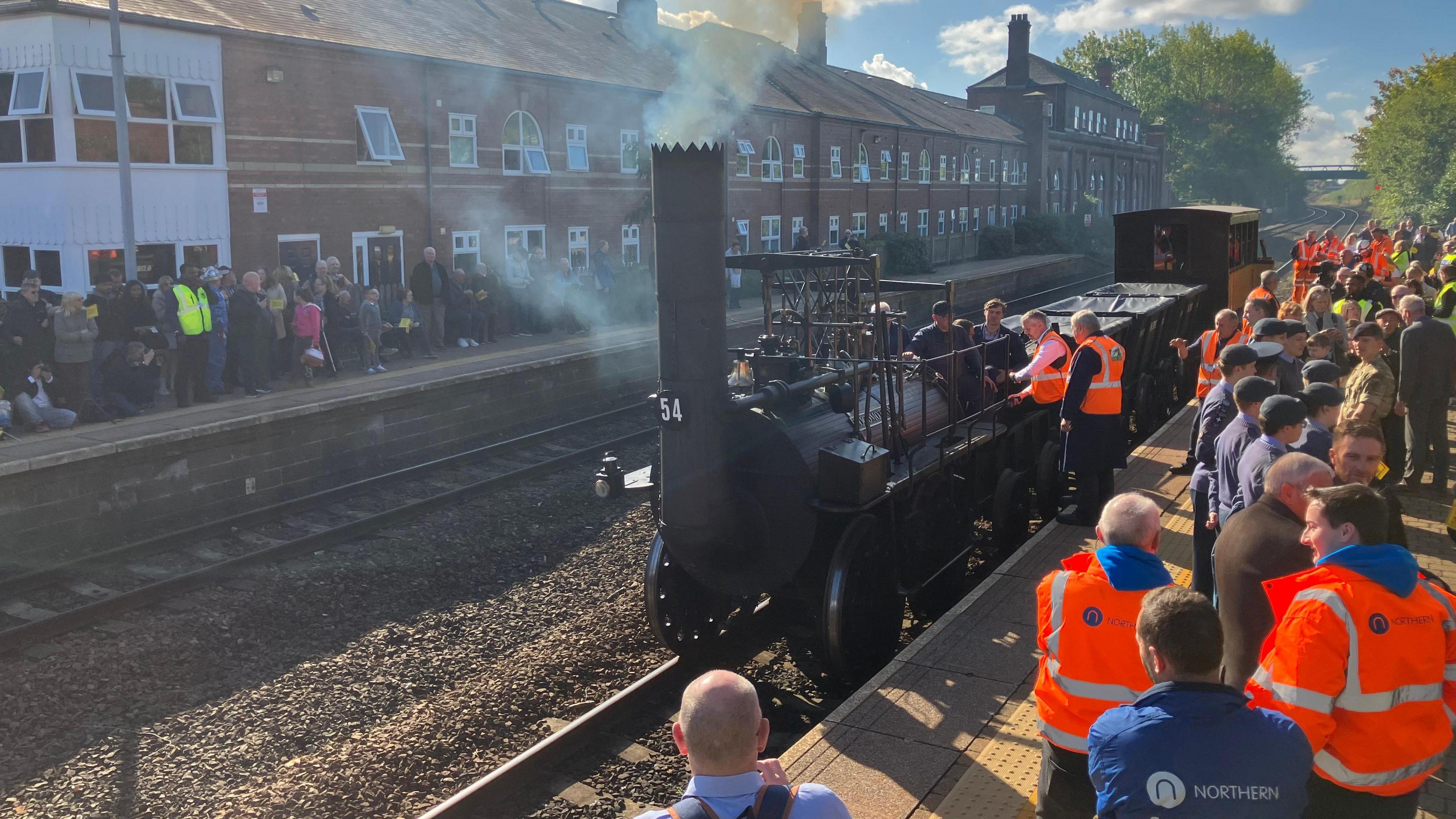 A crowd of people, including multiple wearing in bright orange jackets, stand on two platforms as a black steam engine and train passes though a station