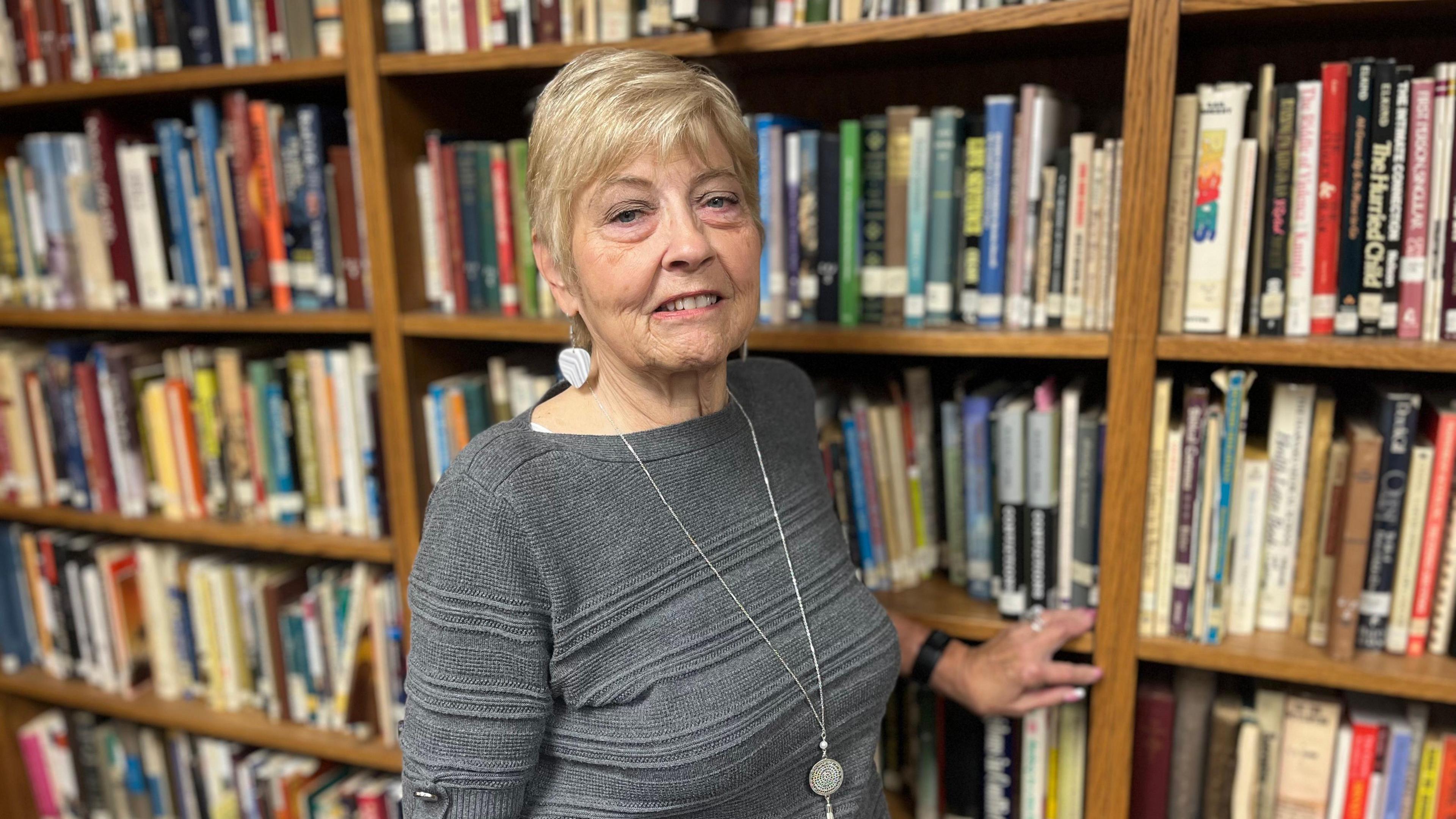 Kathy Magner with short blonde hair standing in front of a bookcase. She is wearing a grey top and a silver necklace.