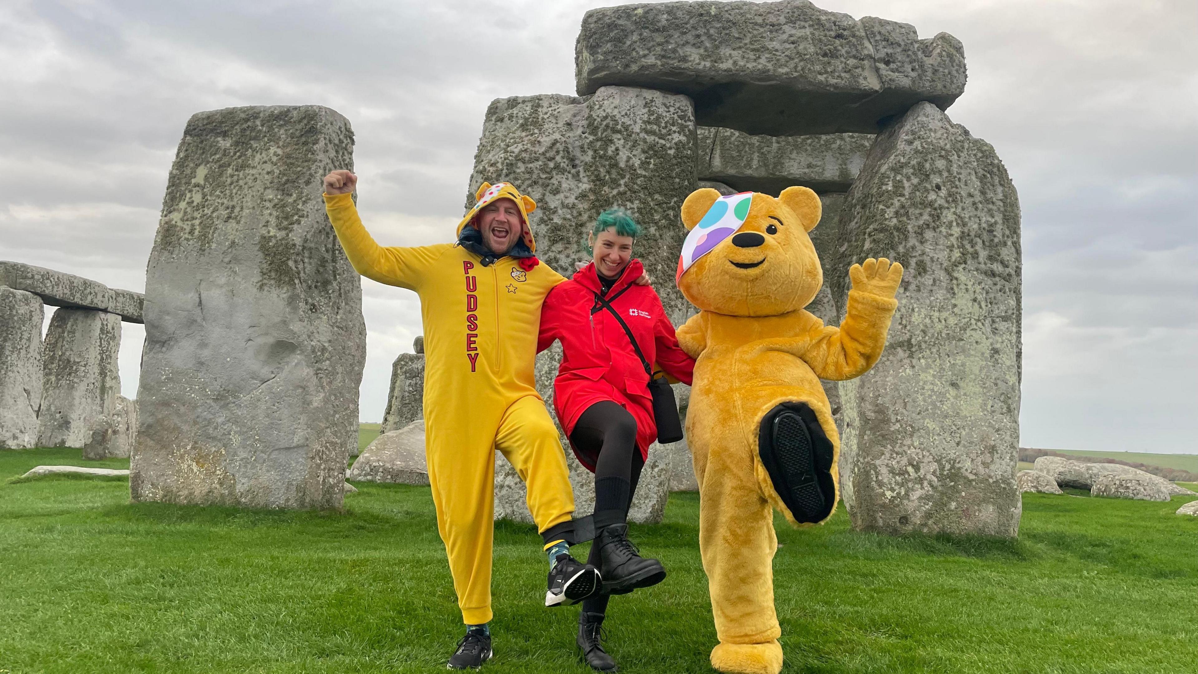 A man wearing a yellow Pudsey Bear onesie has his arm around someone else with blue hair and a red coat, who in turn has their arm around lifesize Pudsey Bear. All three are standing in front of Stonehenge and each have a leg off the floor