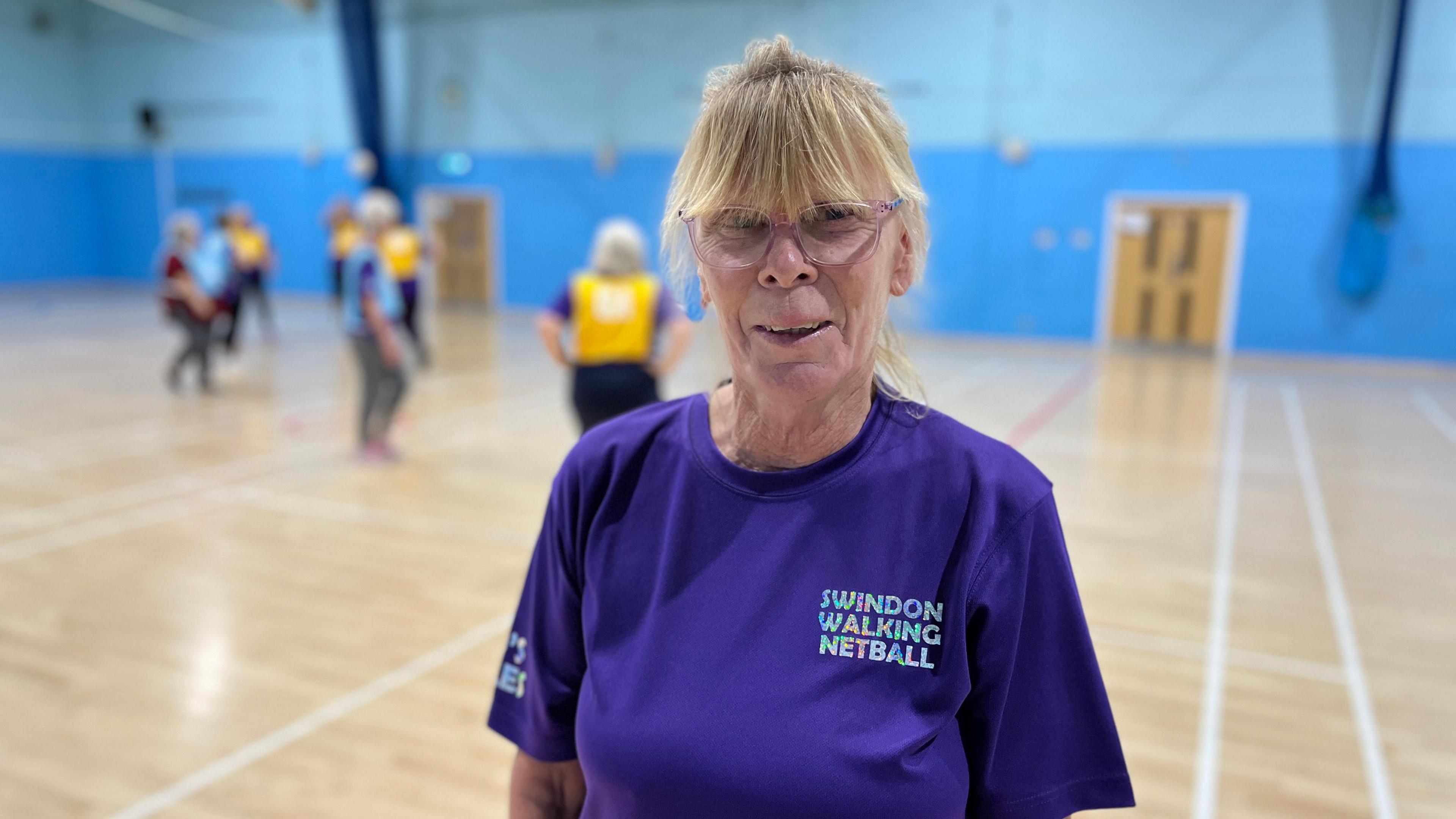 Chris Geddes looks into the camera smiling. She's wearing a Swindon Walking Netball purple t-shirt and glasses. In the background women are playing walking netball.