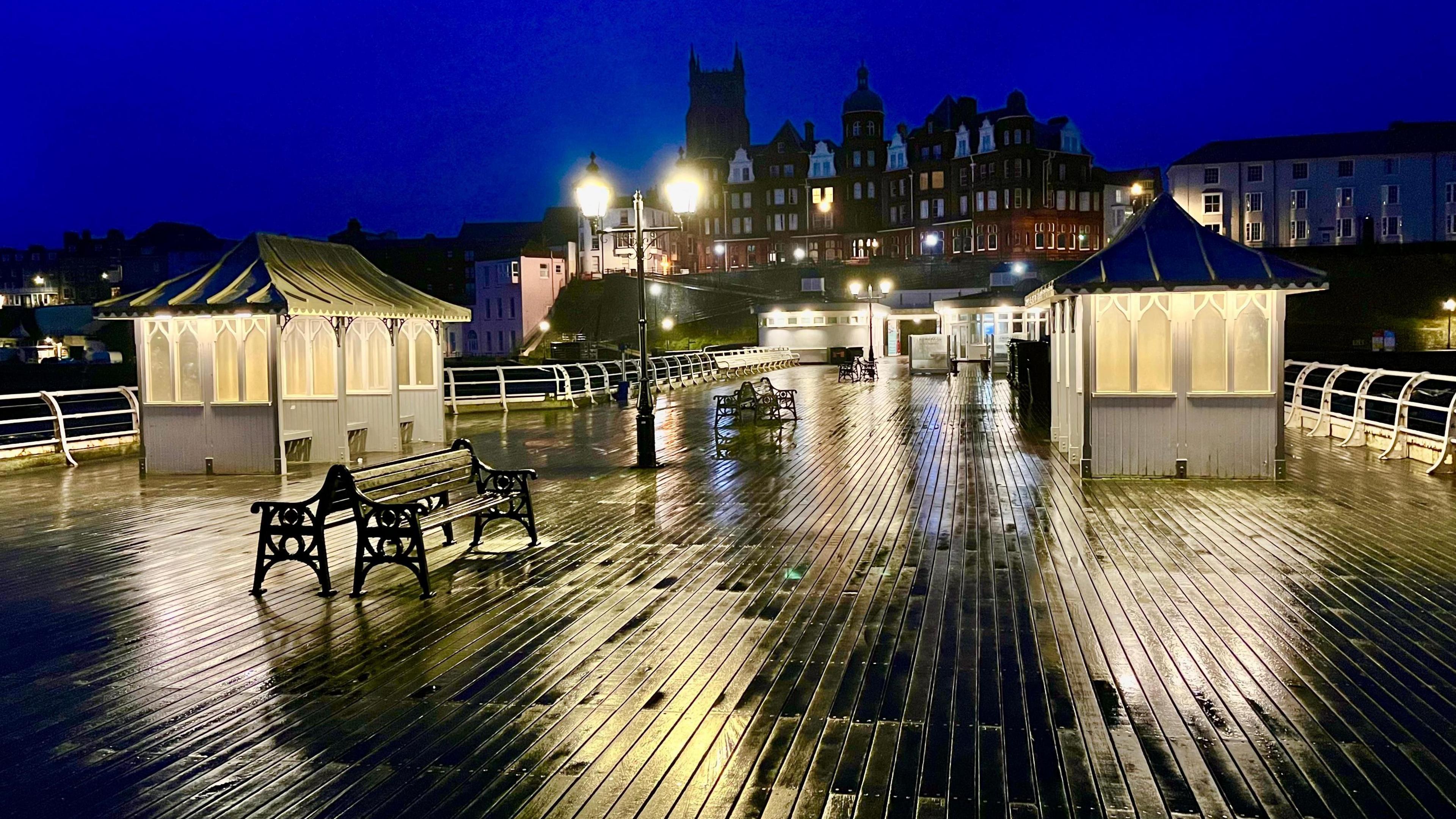 A seaside pier with water on the wooden planks. You can see large buildings in the distance, and a light on the pier, with benches and white shelters. 