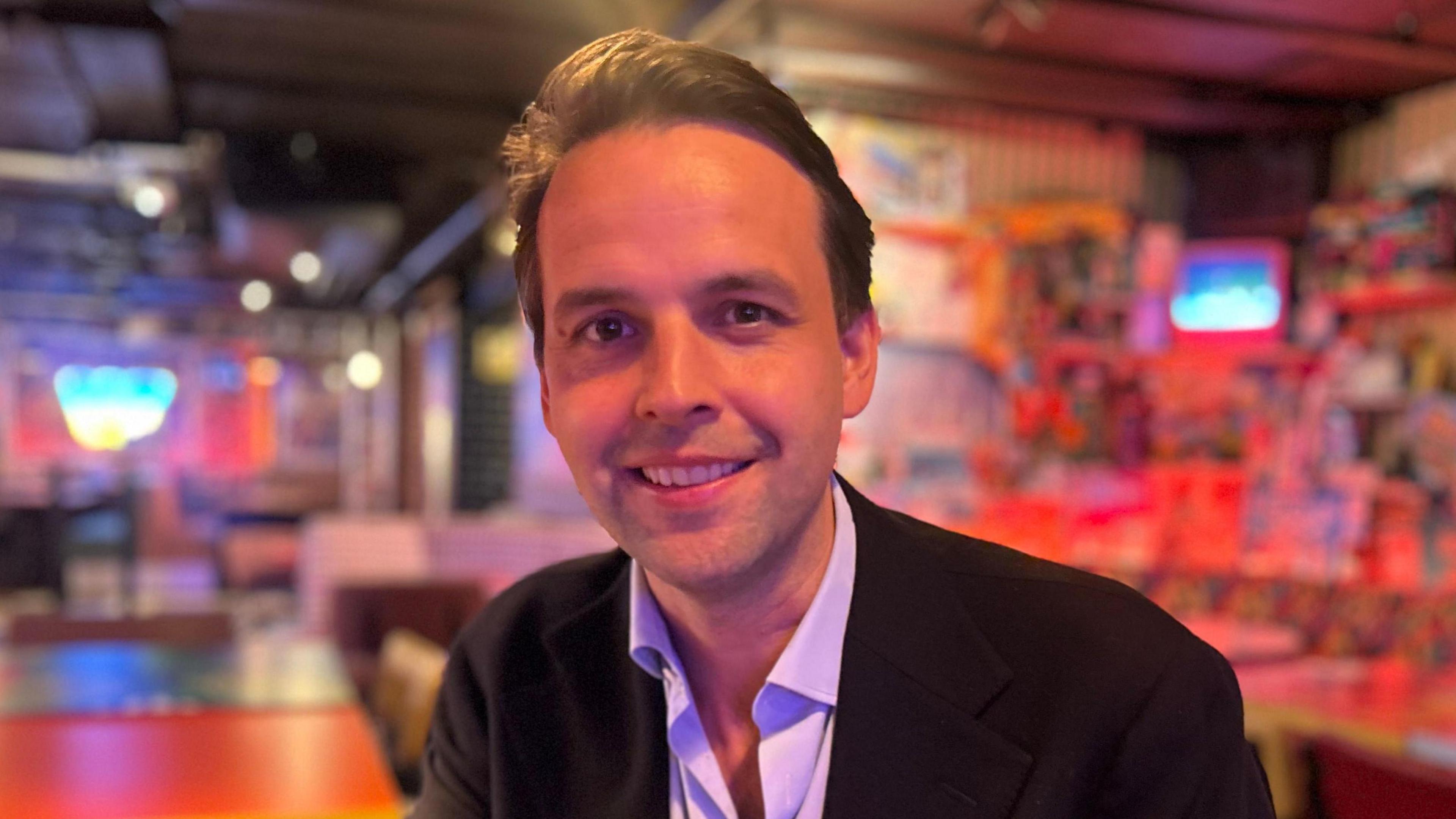 A man with brown hair looks smiling at the camera. He is wearing a dark jacket and a shirt with no tie