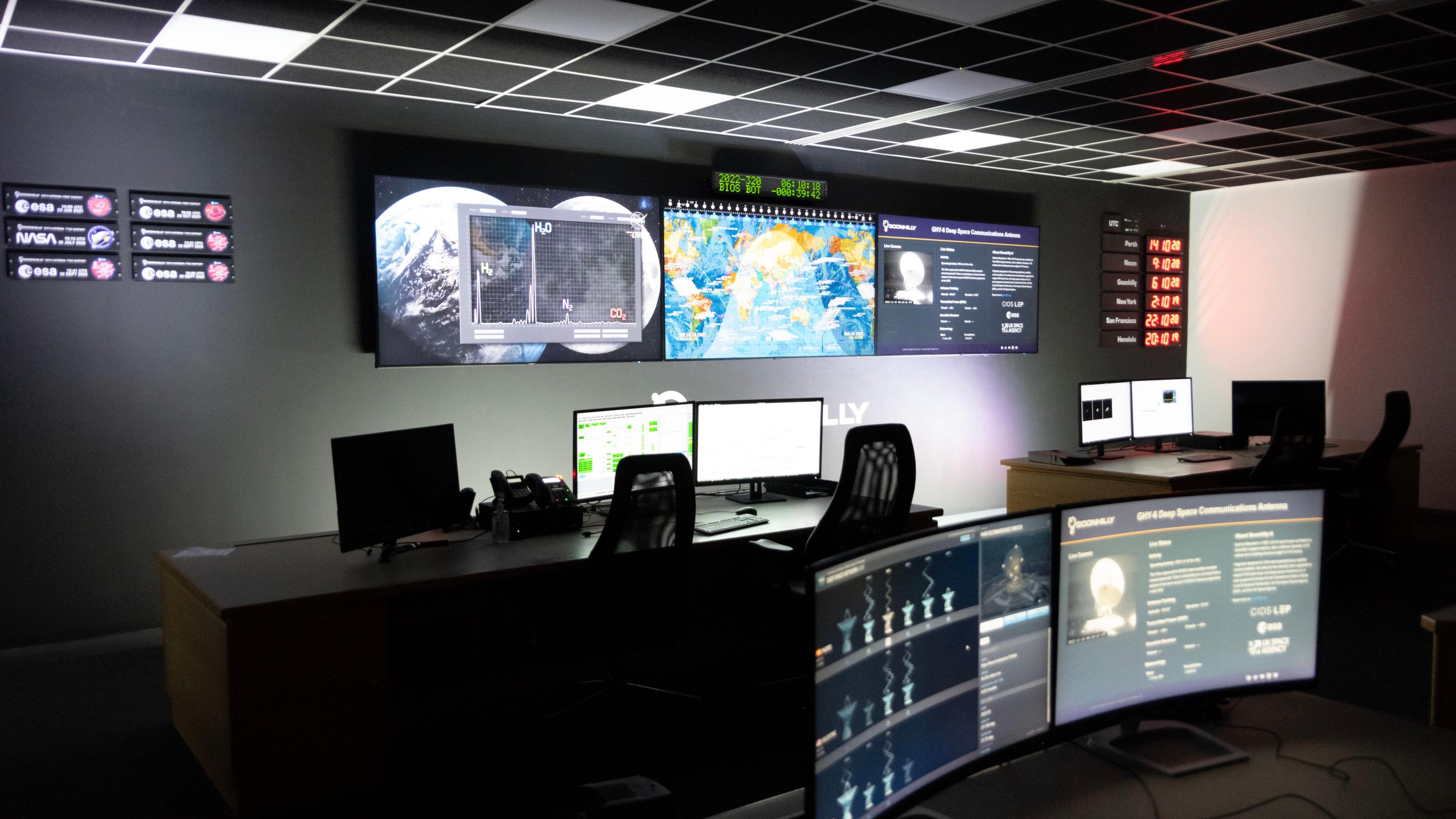 A number of flat screens on a wall show visuals of space with two desks with computer screens and a further two screens behind and a ceiling wtih a chequerboard pattern with an overwhelming aesthetic of space exploration.