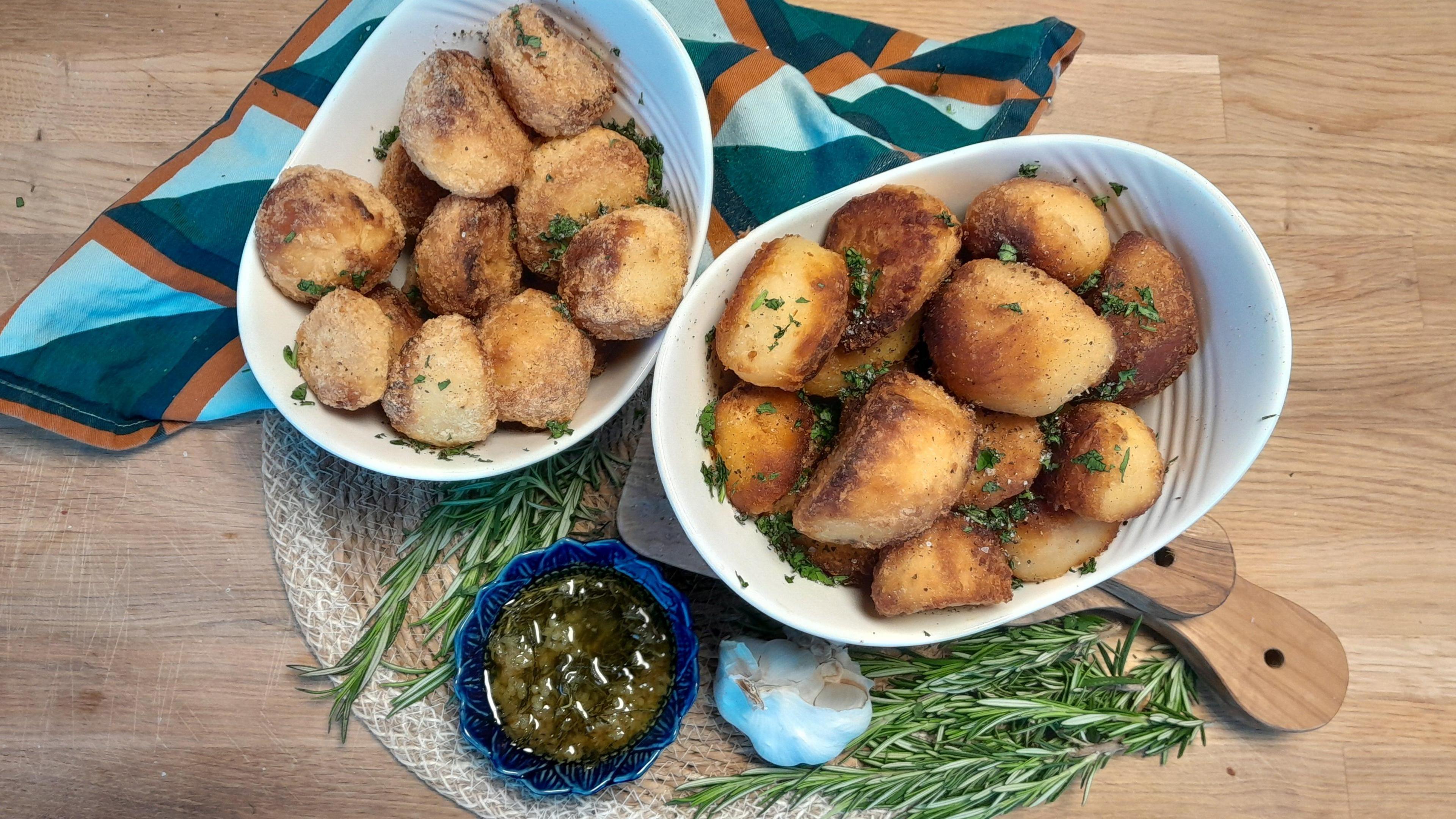 Two white ceramic bowls of Yvonne's golden-brown roast potatoes, scattered with green herbs. The bowls are sitting on a wicker mat on a wooden board, with a blue and orange napkin at the side. A small blue dish contains a serving of dark green rosemary and garlic butter. A bulb of garlic and some sprigs of rosemary are scattered around the edges.