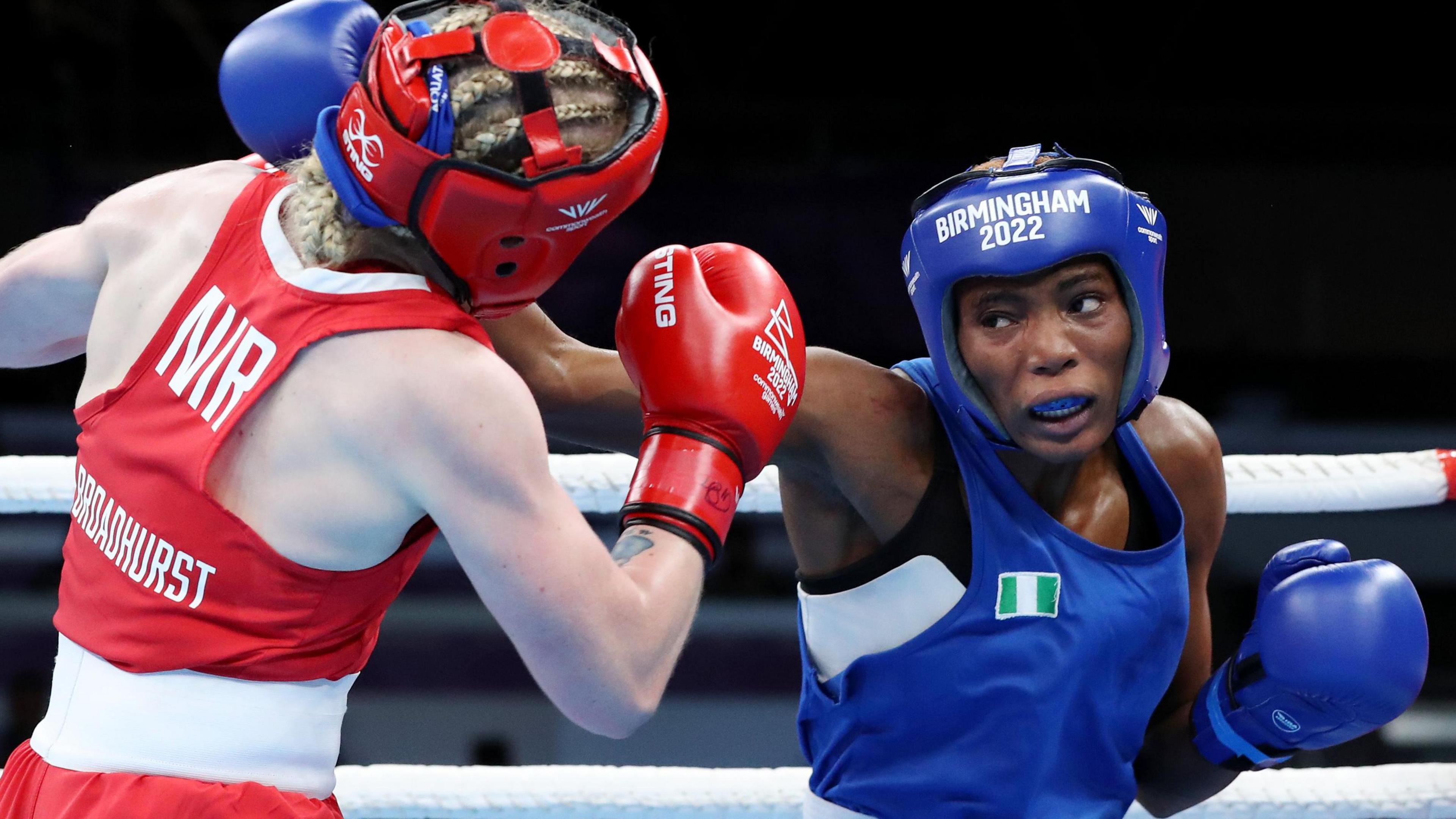 Nigerian boxer Cynthia Ogunsemilore, wearing blue vest, headguard, gloves and gumshield, lands a right-handed punch on an opponent dressed in red in the ring