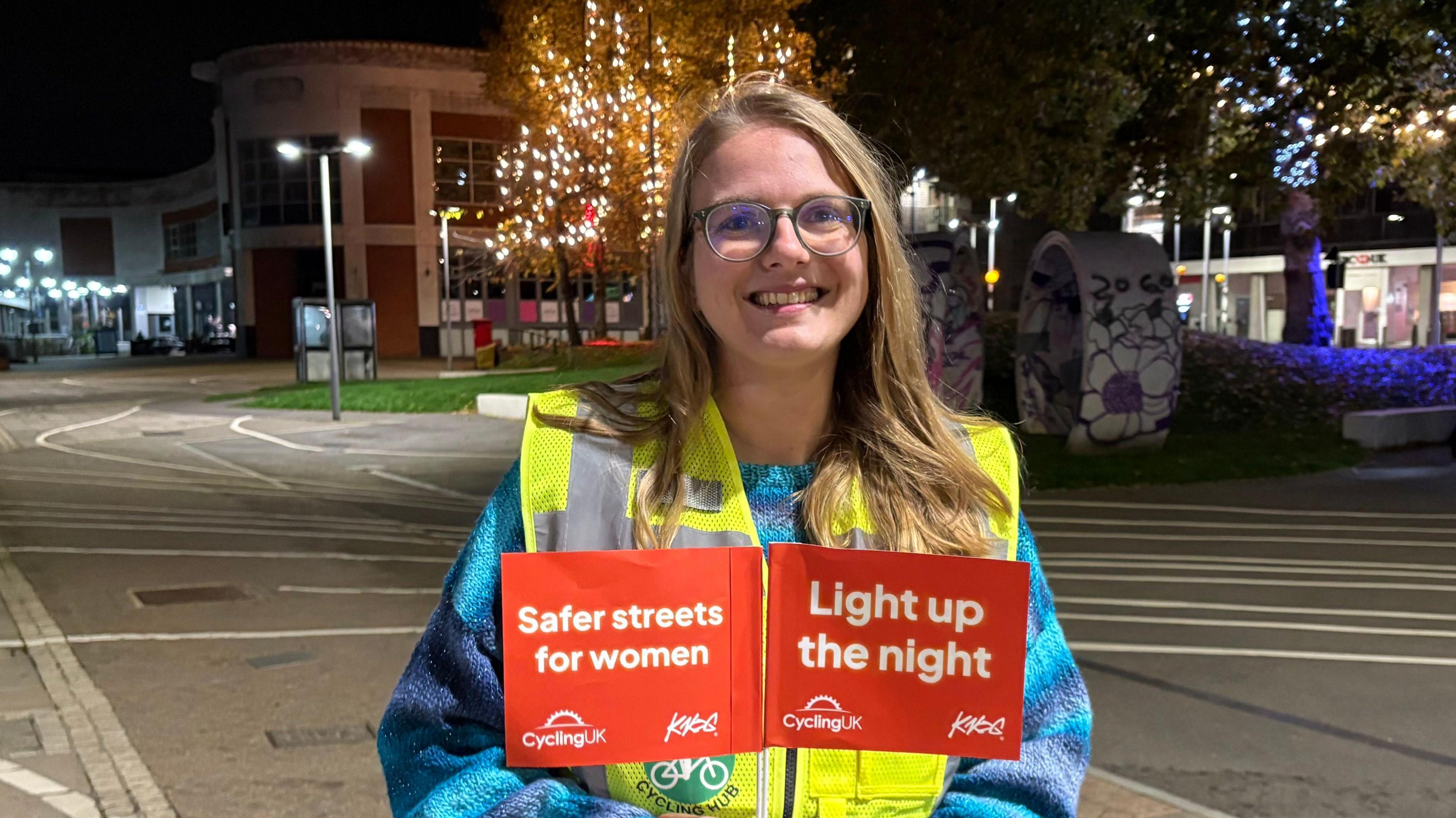 Jennifer Huygen with long blond hair and glasses smiling at the camera and holding up small orange flags with the words "Safer Streets for Women" and "Light up the Night" in white writing. She is wearing a yellow hi-vis vest and blue coat and is standing in front of a large pedestrianised area with trees with lighted-up branches in the background.
