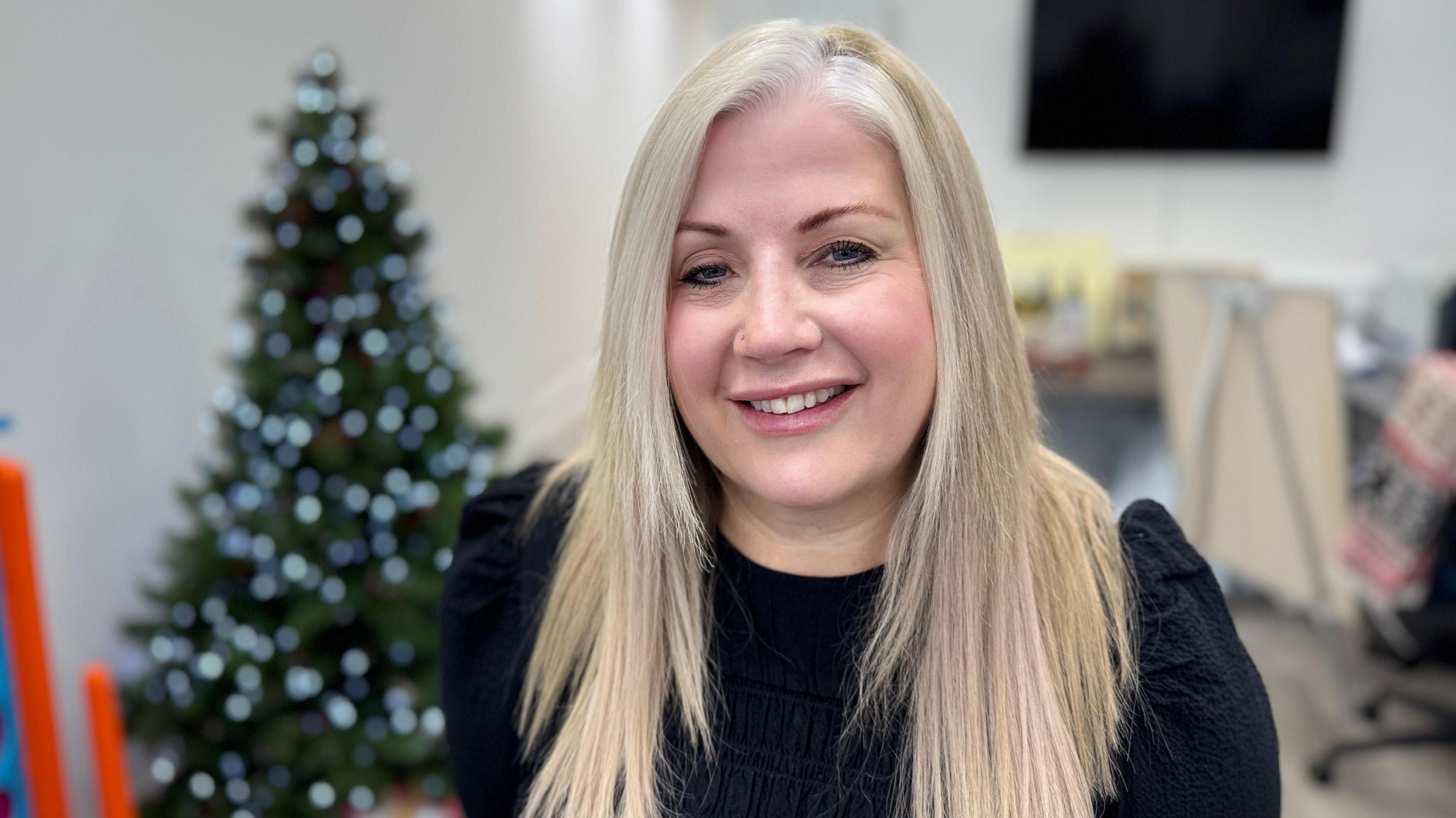 Karen Jones, with blonde hair, is sitting in front of a Christmas tree in her Cardiff office, wearing a black top 