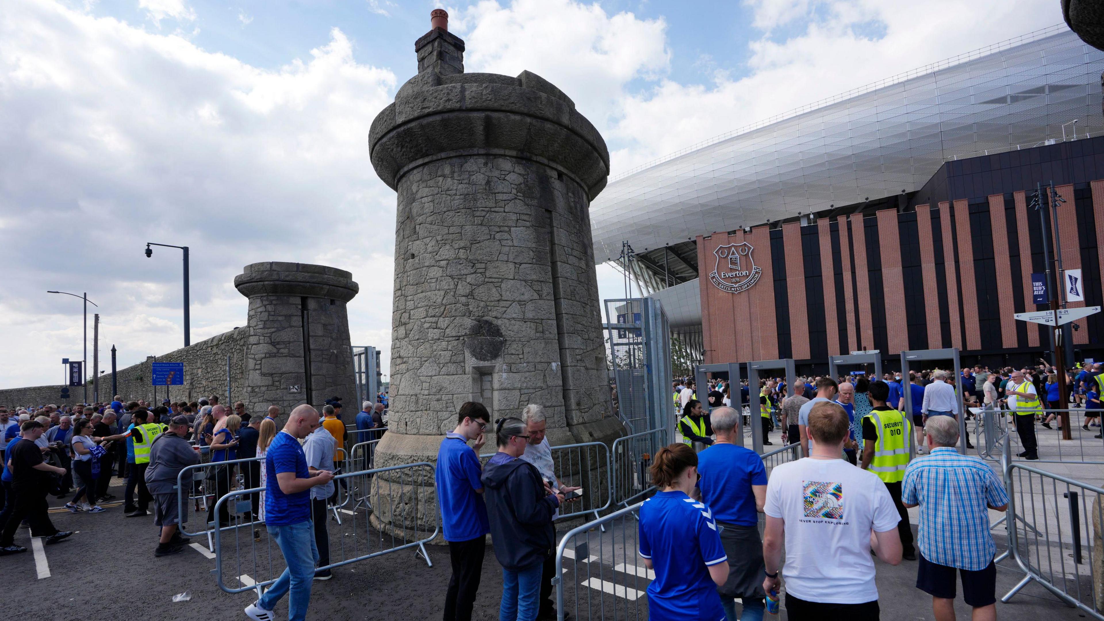 Crowds of fans, mainly in blue clothing, line up in queues leading to security gates that are next to tall dock towers made of grey granite stone. The old dock wall continues around the stadium behind. The arena has a striped orange and black front with the Everton logo under a silver metal roof.