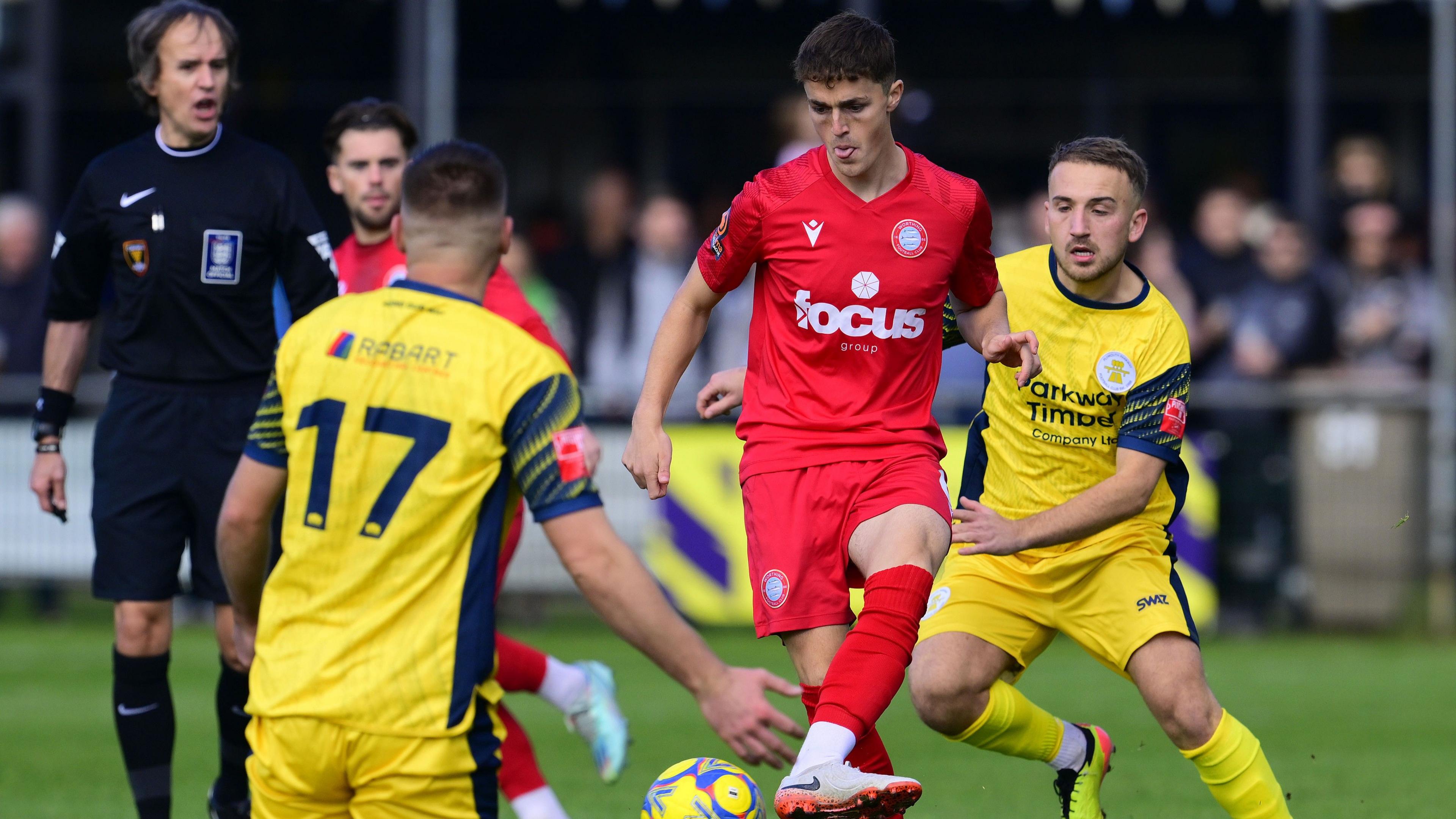 Jack Wadham passes the ball with his instep in a crowded midfield, wearing Worthing's all red home kit