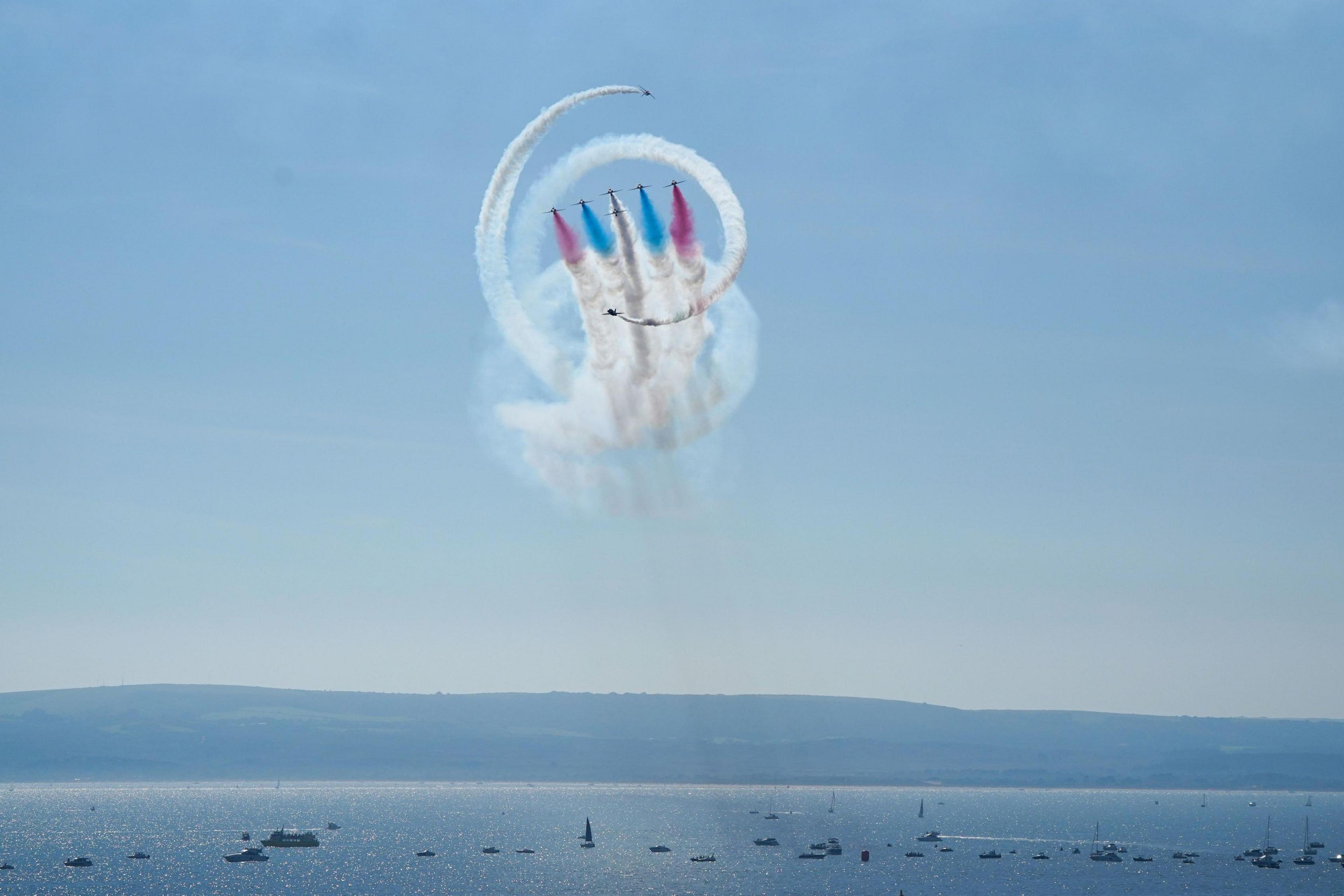 The Red Arrows performing the Tornado over the glittering sea at Bournemouth. The manoeuvre involves six aircraft flying towards the shore in formation while creating red and blue smoke trails while two other aircraft fly in a spiral around them them creating a double spiral trail of white smoke around the blue and red trails.