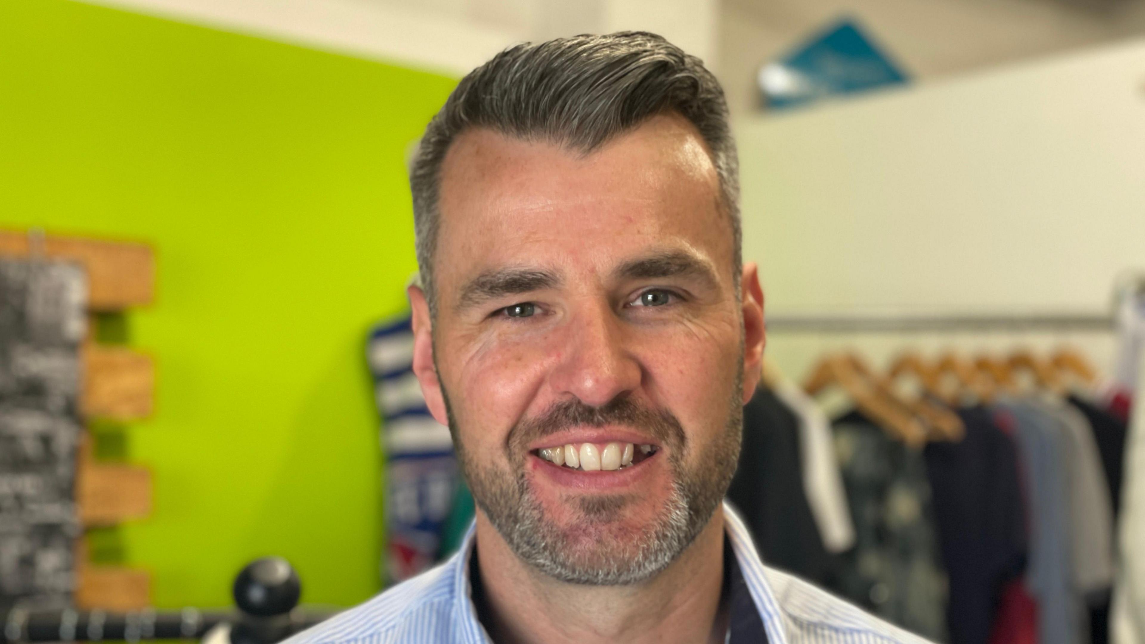 A close up of Oliver Miller, from Bishop & Miller Auctioneers. He is smiling directly into the camera. A row of tops on hangers can be seen in the background on a clothes rail.