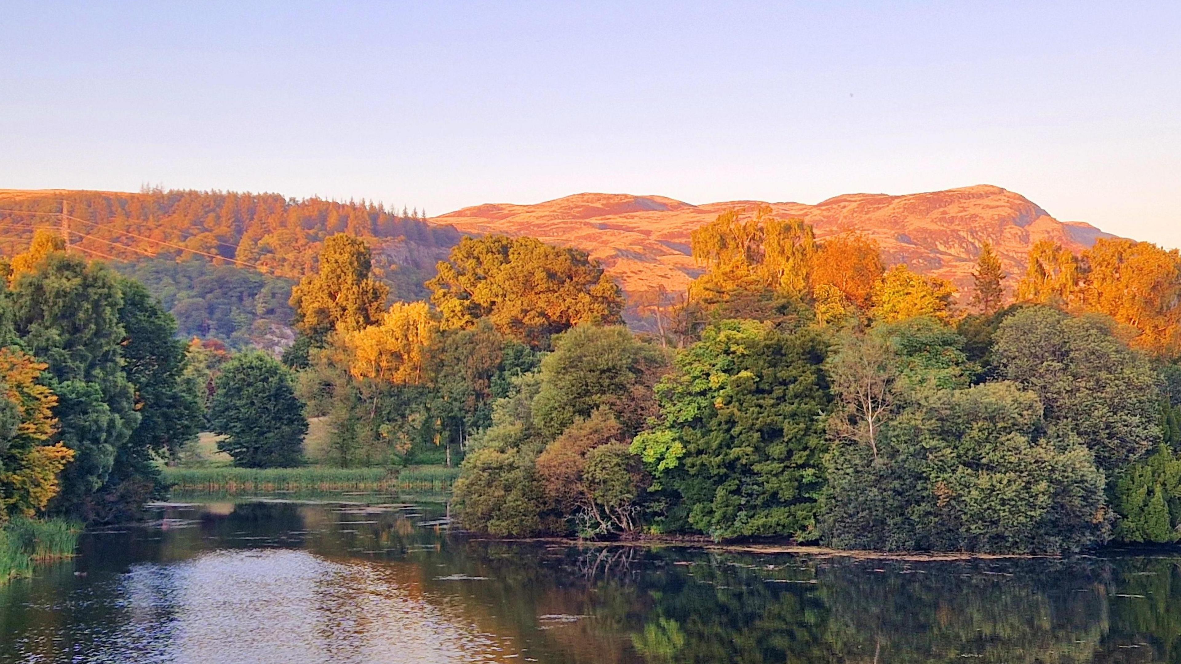 View across a lake with green trees surrounding and behind, some trees that have started to turn golden brown. In there background there are hills lit golden brown by the setting Sun