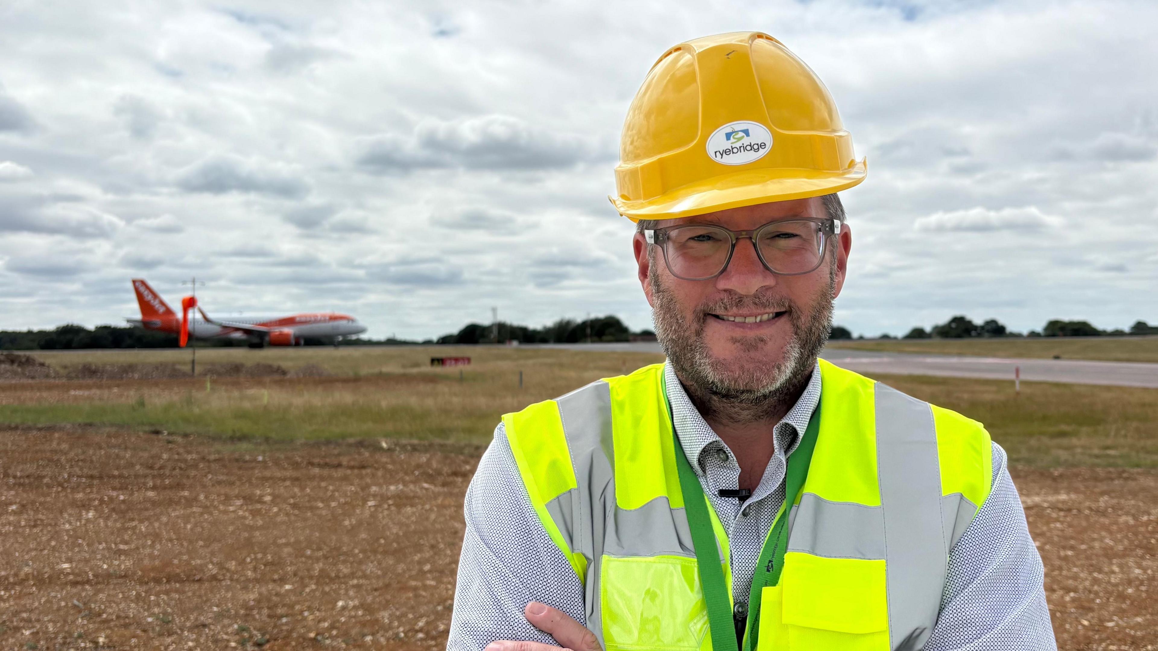 Sean Scully smiling and looking direct to camera. He is wearing a yellow hard hat and a green florescent jacket, over a checked shirt. He has a stubbly beard and glasses. He is standing outside and in the background is an orange and white plane that is either landing or taking off on a runway. It is a bright day but there are many clouds in the sky.
