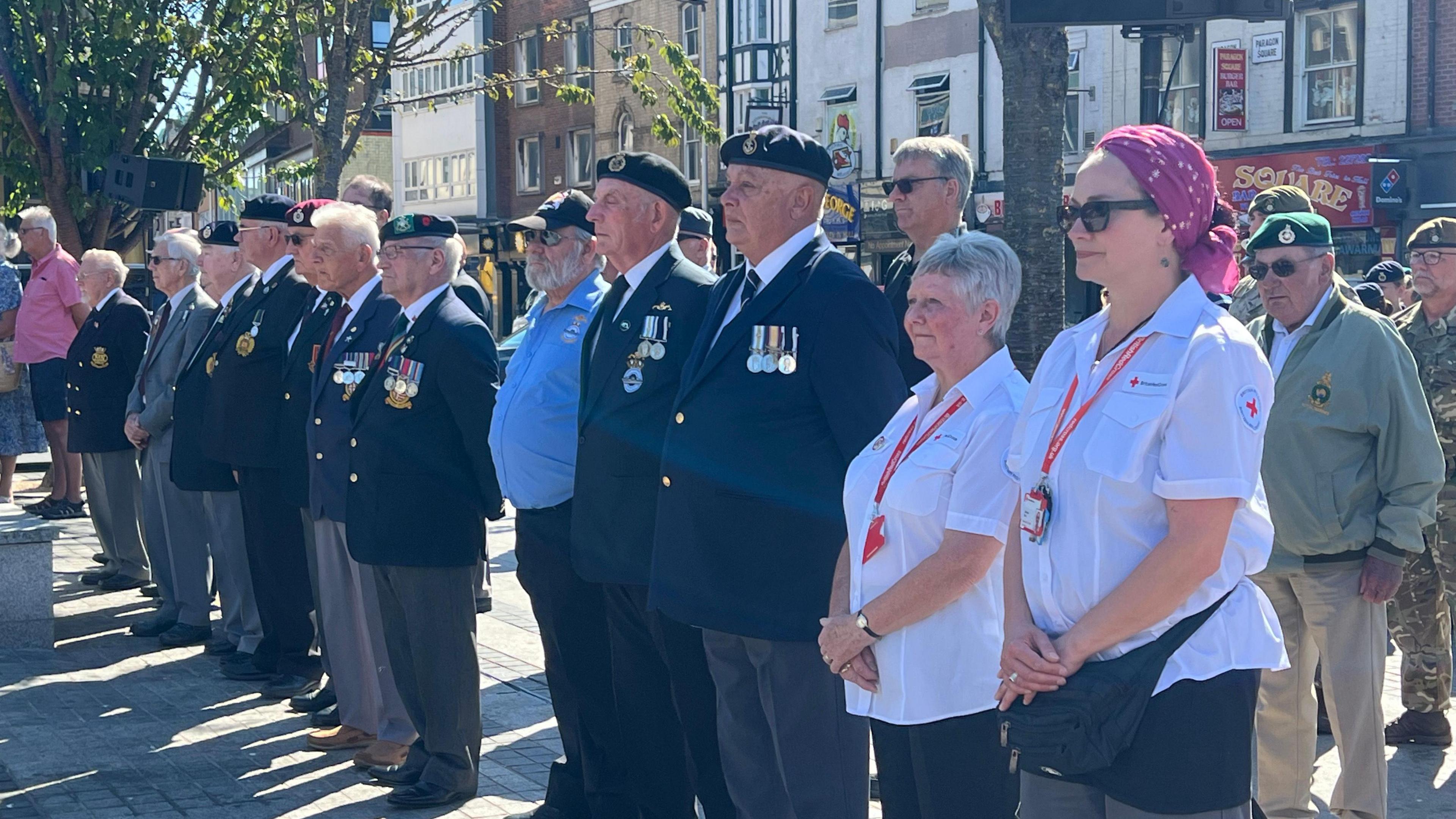 A group of people standing in a line outside. The people on the left are mostly men in dark blazers with medals. The people on the right include two women in white shirts. There are buildings in the background.
