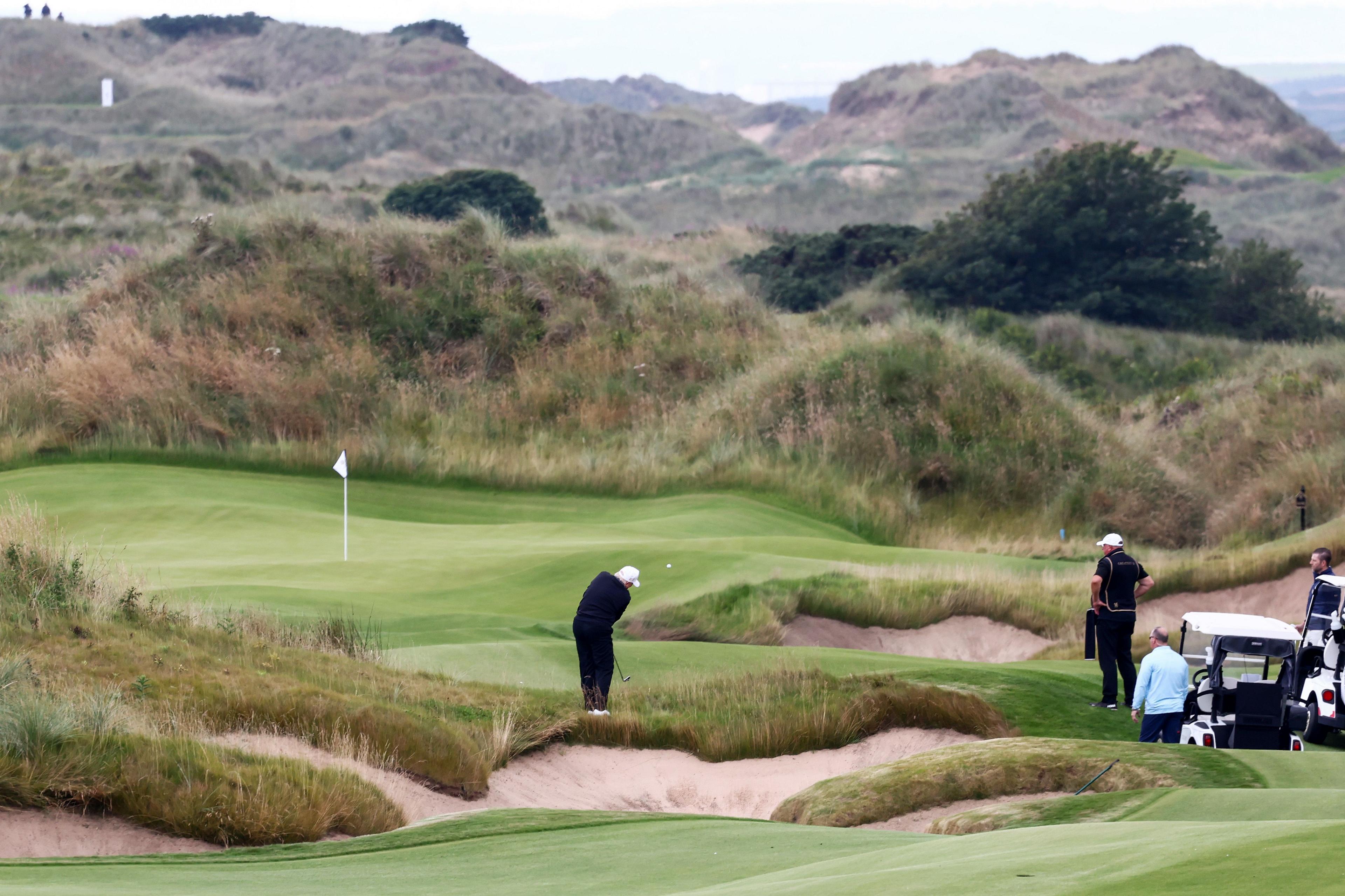Distant photograph of President Donald Trump playing an approach shot to a perfectly manicured green during a round of golf at his new golf course in Balmedie. He is being watched by three men gathered round a golf buggy on the right of the image. Rolling sand dunes can be seen in the distance. Trump - who is a tiny figure in the scenic shot - is dressed in black and is wearing a white baseball cap and white golf shoes. 