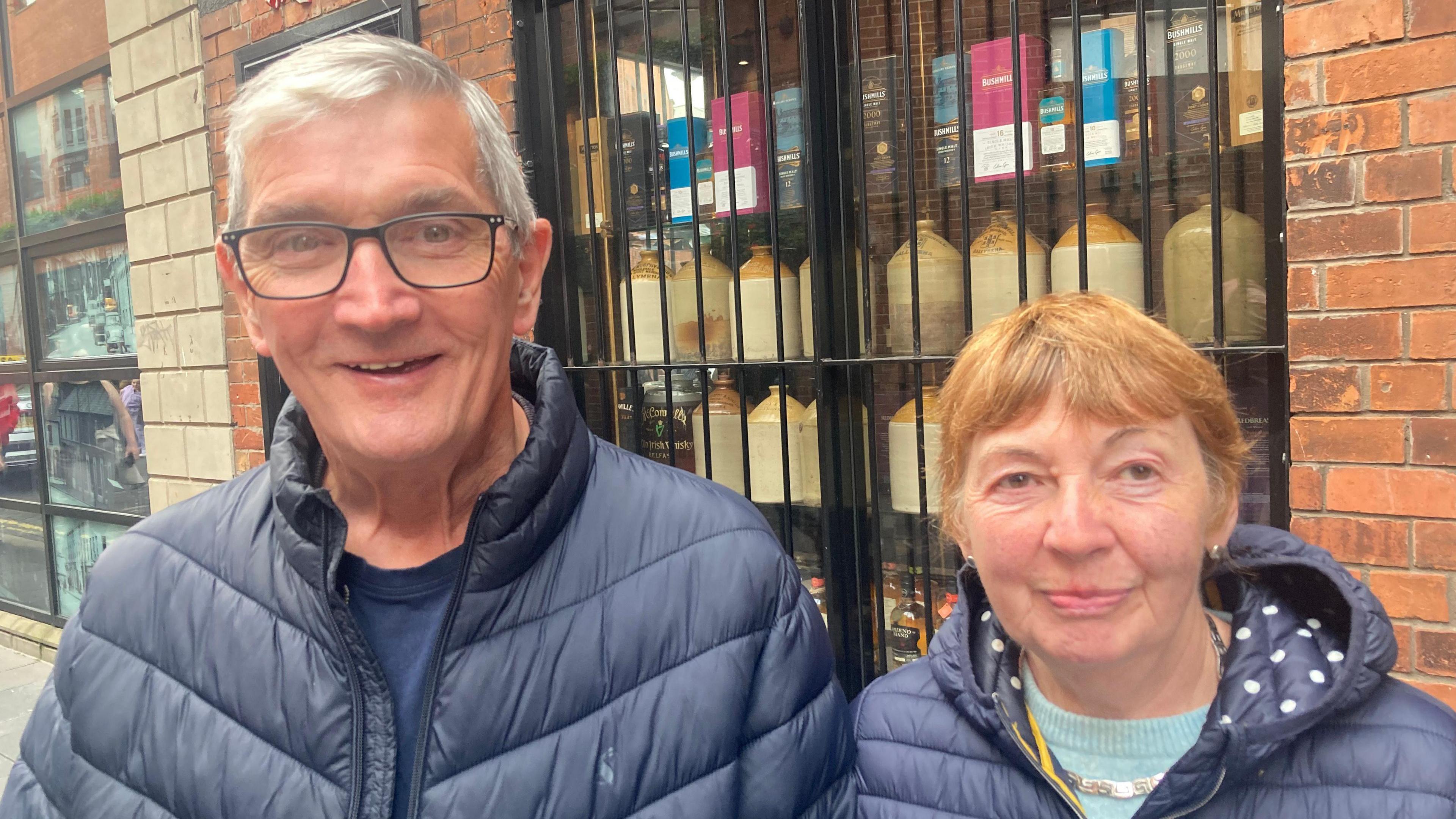 Tom and Roisin Martin standing on a street in Belfast city centre.  They are both wearing navy padded jackets.  Tom Martin has short grey hair and glasses.  Roisin Martin has short, reddish hair and is wearing a pale blue jumper under her coat.  Bottles of whiskey and jars are displayed in the window behind them. 