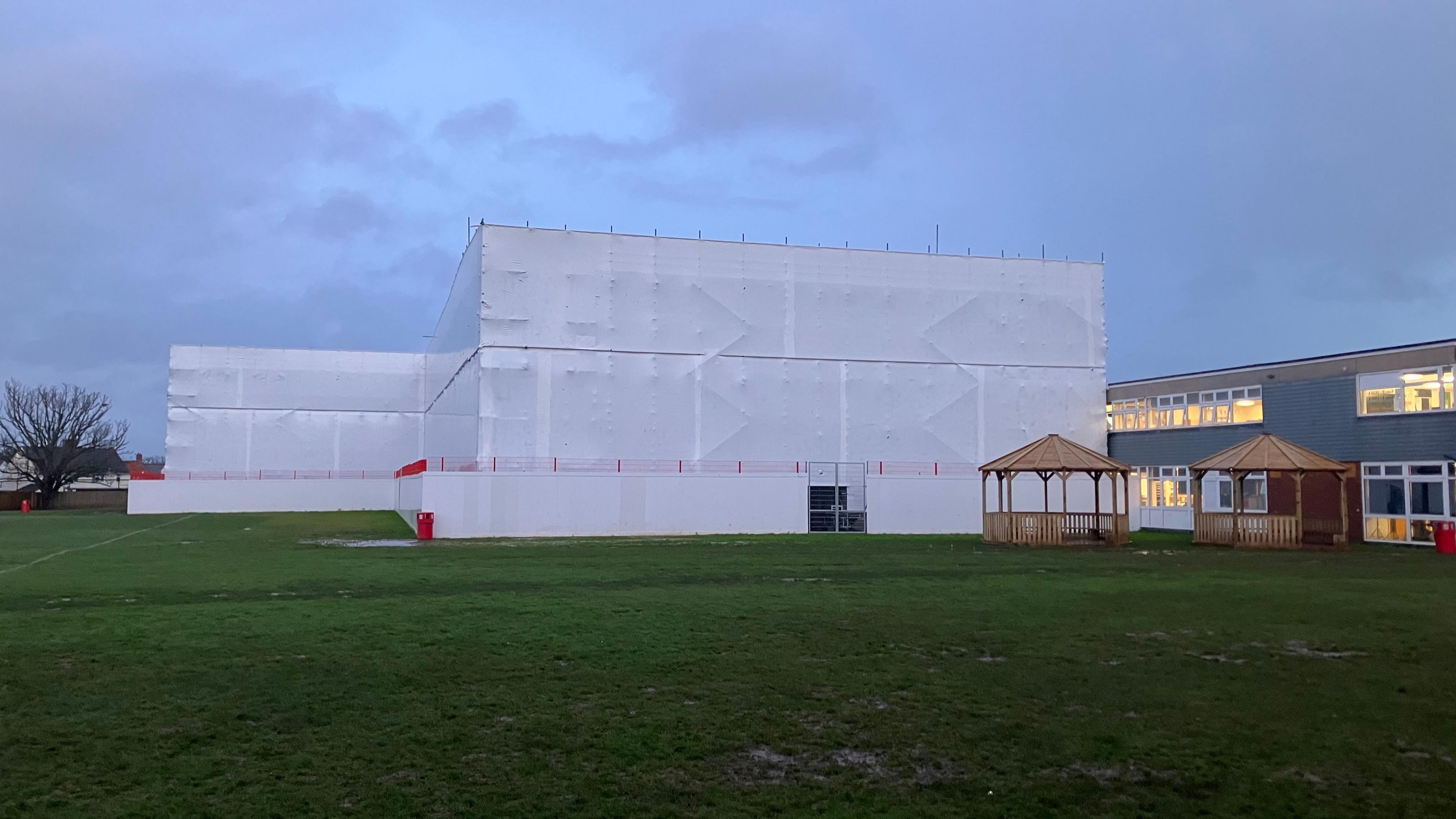 White sheeting covered scaffolding around a school building in a playing field at dusk with the lights on in some neighbouring classroom buildings and two wooden gazebos on the field