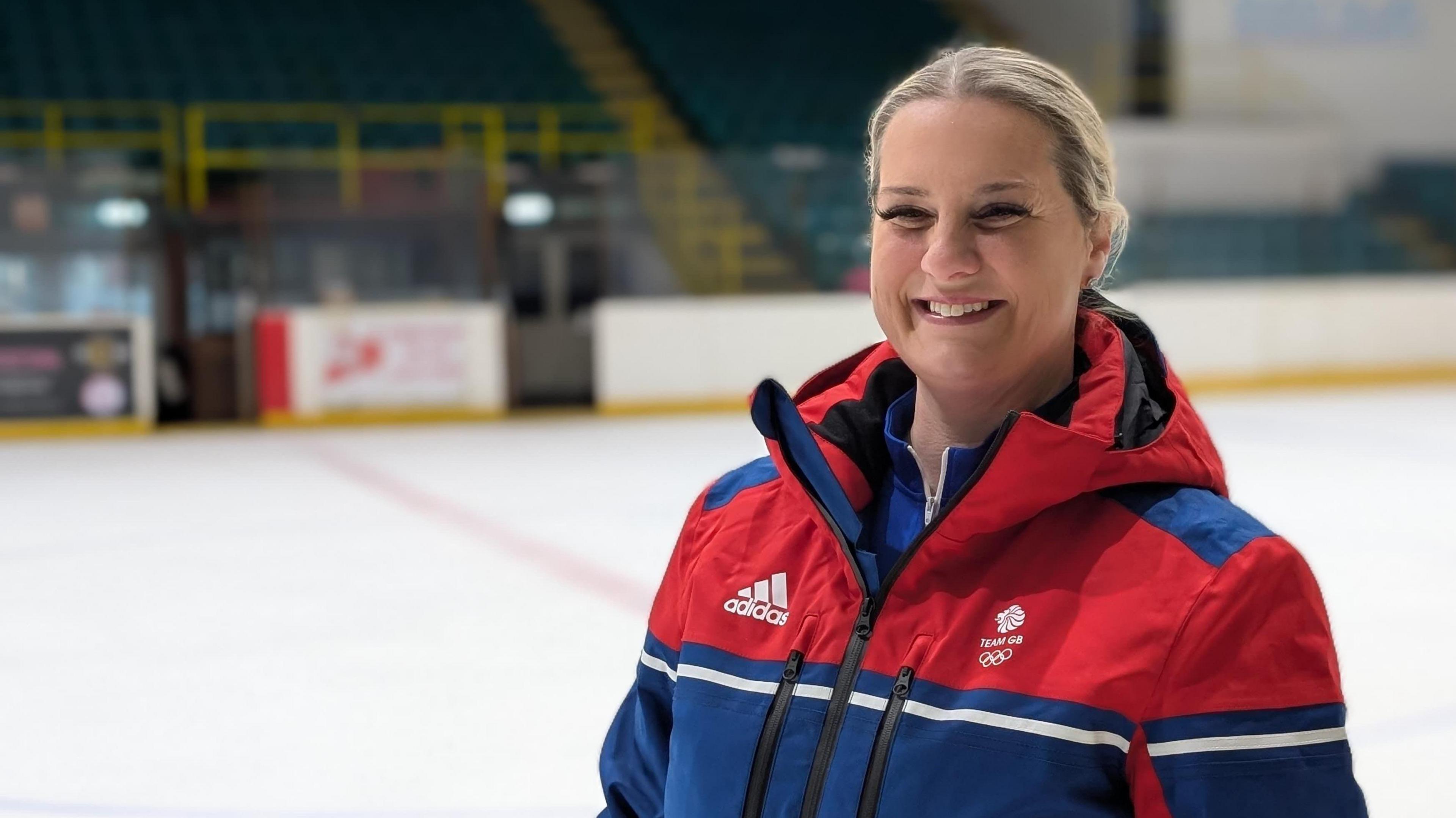 Karen Brennan wears a red white and blue Team GB coat and is smiling. The ice and stands of Whitley Bay Ice Rink are blurred out in the background. 