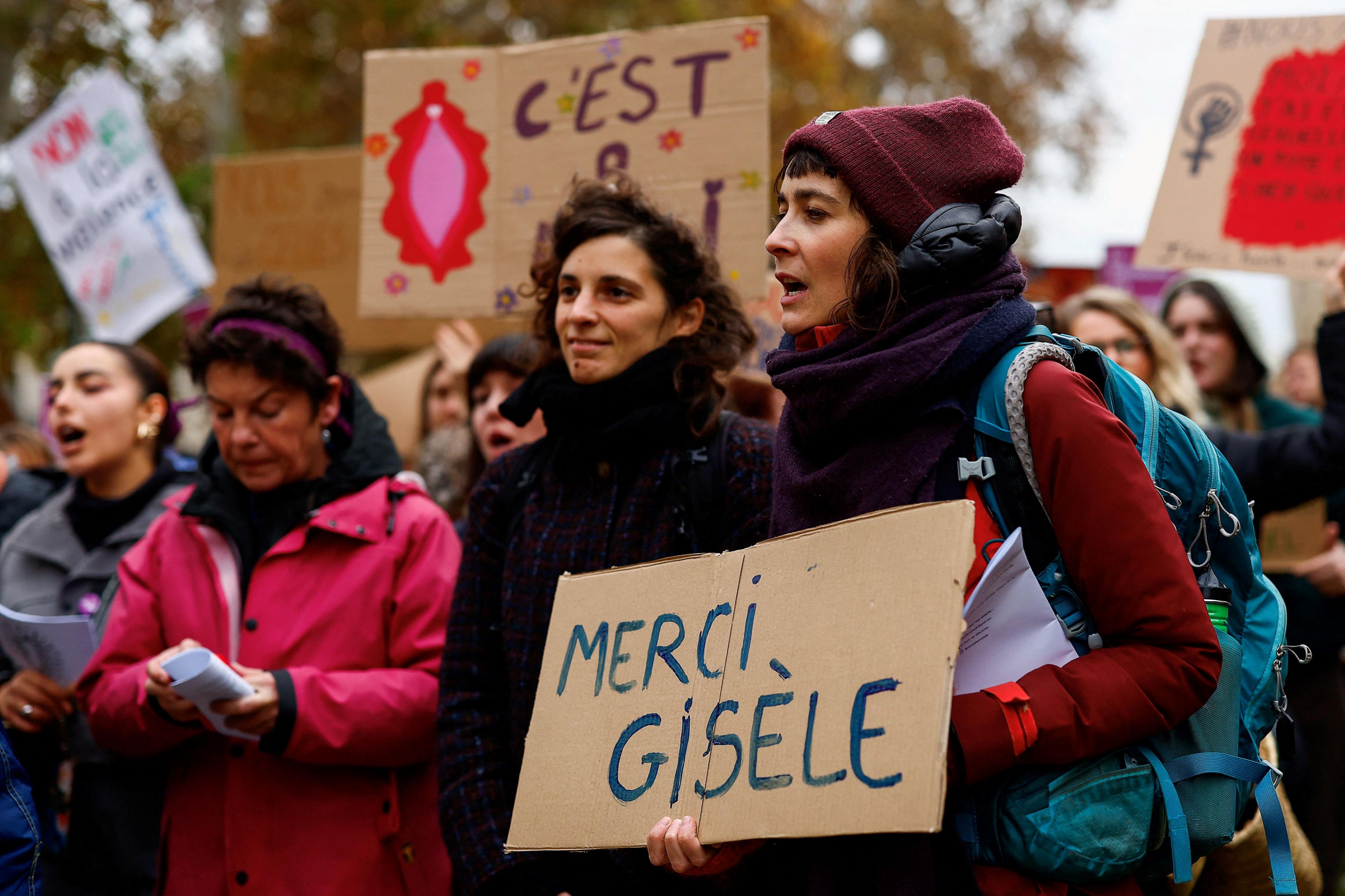 A group of women stand outside, holding placards. One woman at the front holds a cardboard sign that reads "Merci Gisèle"