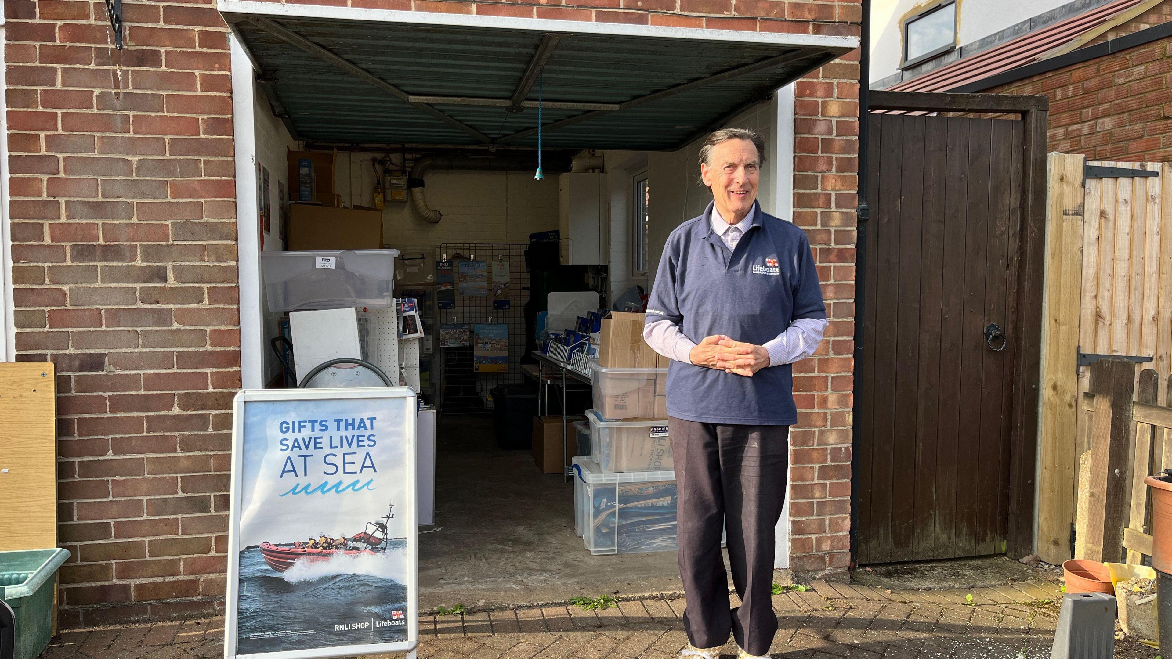 Mr Heathcock is stood outside the shop, which is brick garage next to a house and a garden gate. The door lift up and there is an outdoor sign that reads 'gifts that save lives at sea'. The space is small and not much taller than Mr Heathcock. 