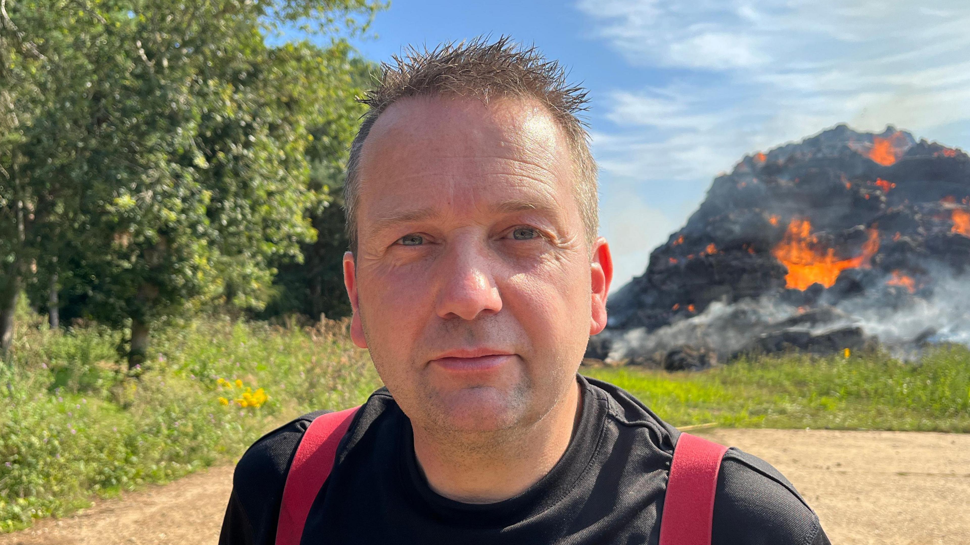 Dave Ellard smiles at the camera in front of the burning stack of hay. He has short brown hair and wears a black T-shirt with red braces over his shoulders. 