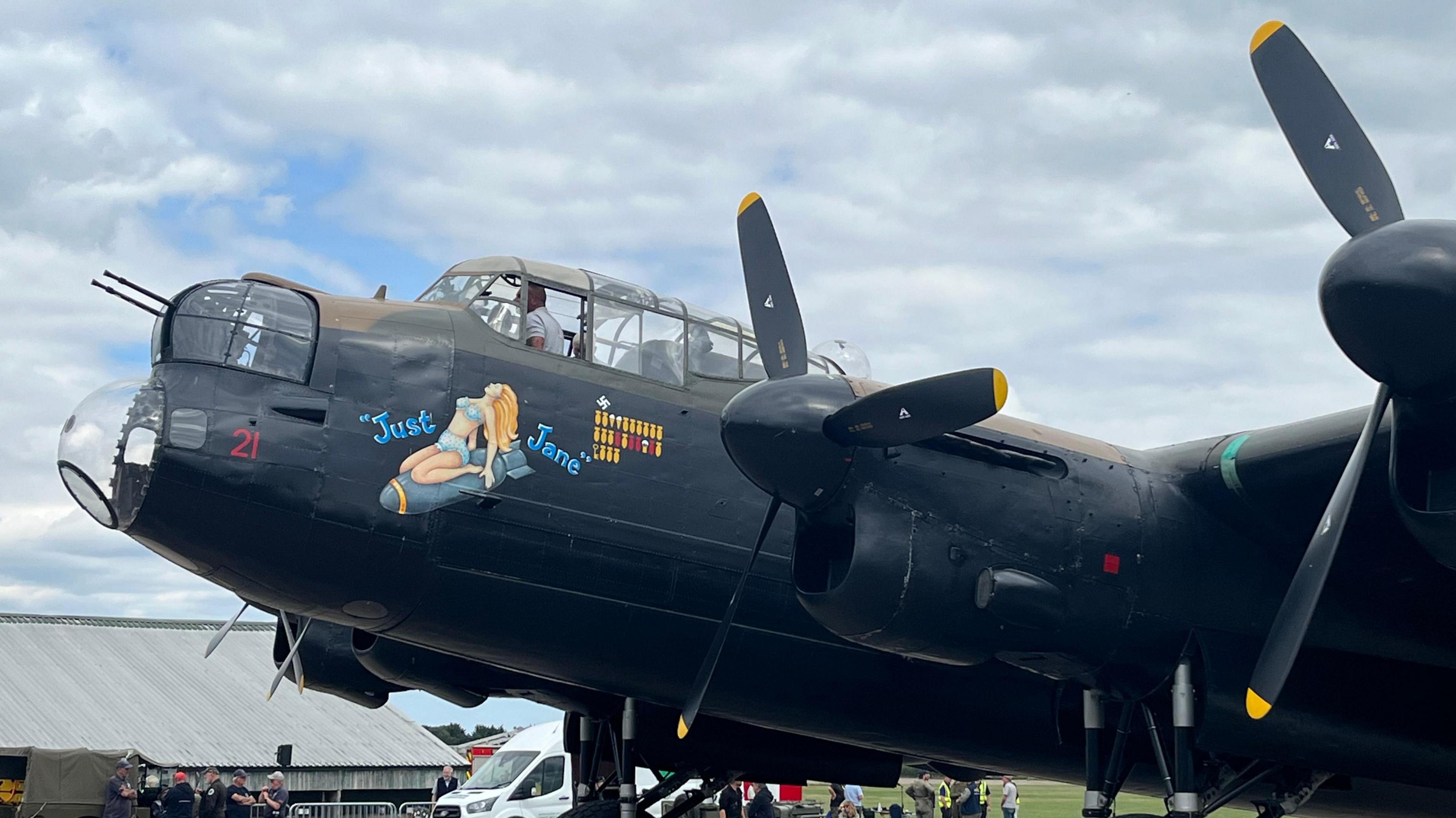 A side view of the nose of a black Lancaster Bomber with an image of a woman in a blue two-piece between blue writing reading "Just Jane".