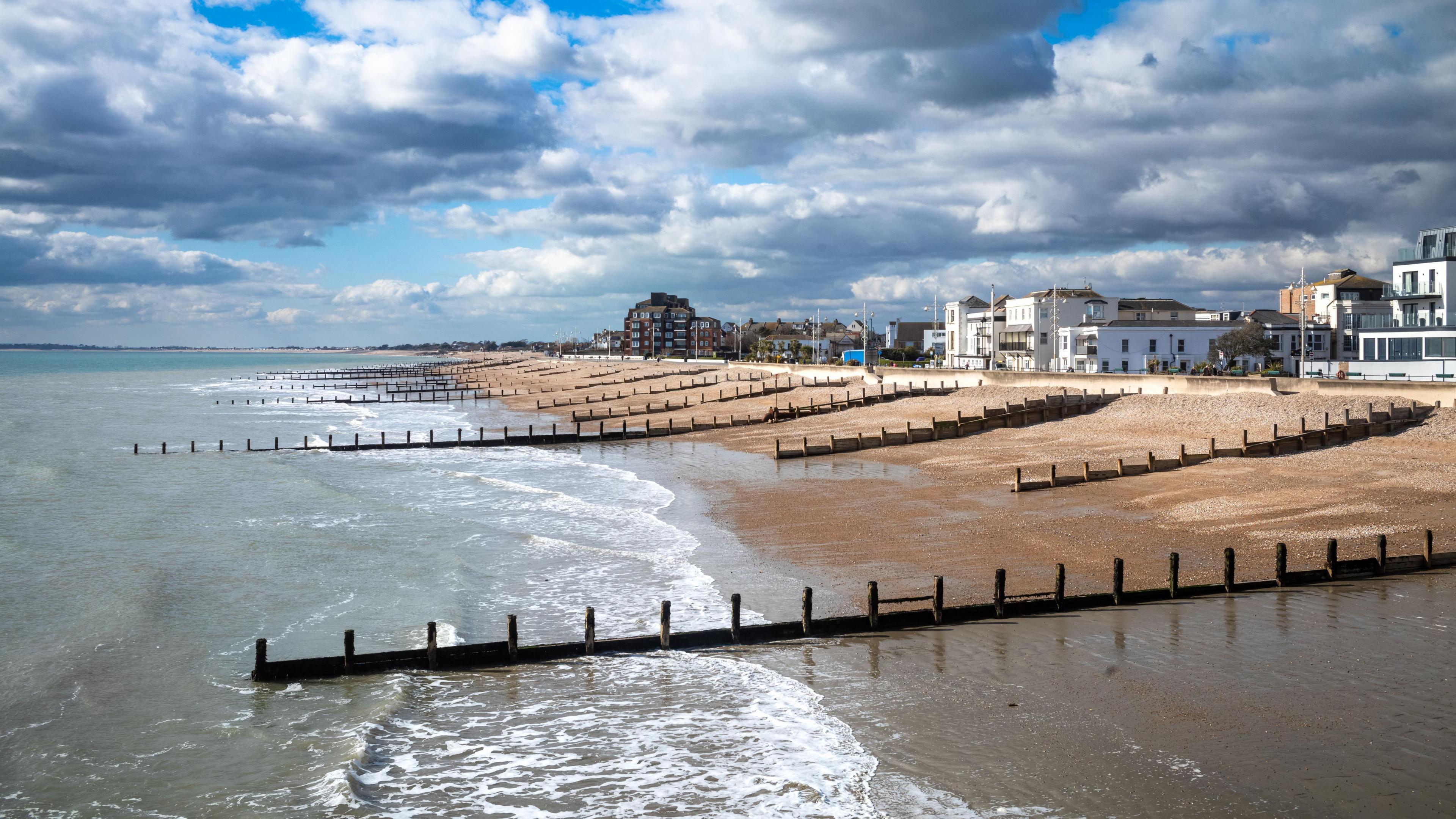 A landscape view of the beach and seafront in Bognor Regis in February. We see the sea in the foreground, the pebble beach with wooden groynes, the seafront buildings and the cloudy sky behind. 