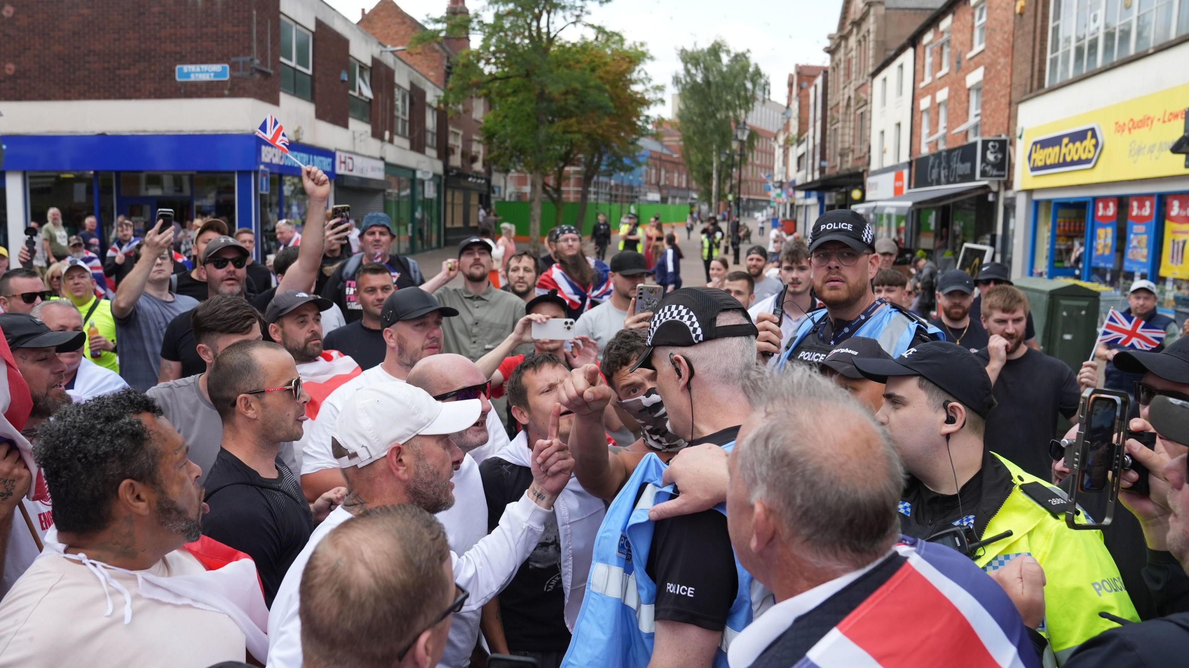 A police officer wearing a blue jacked surrounded by protesters in a town centre