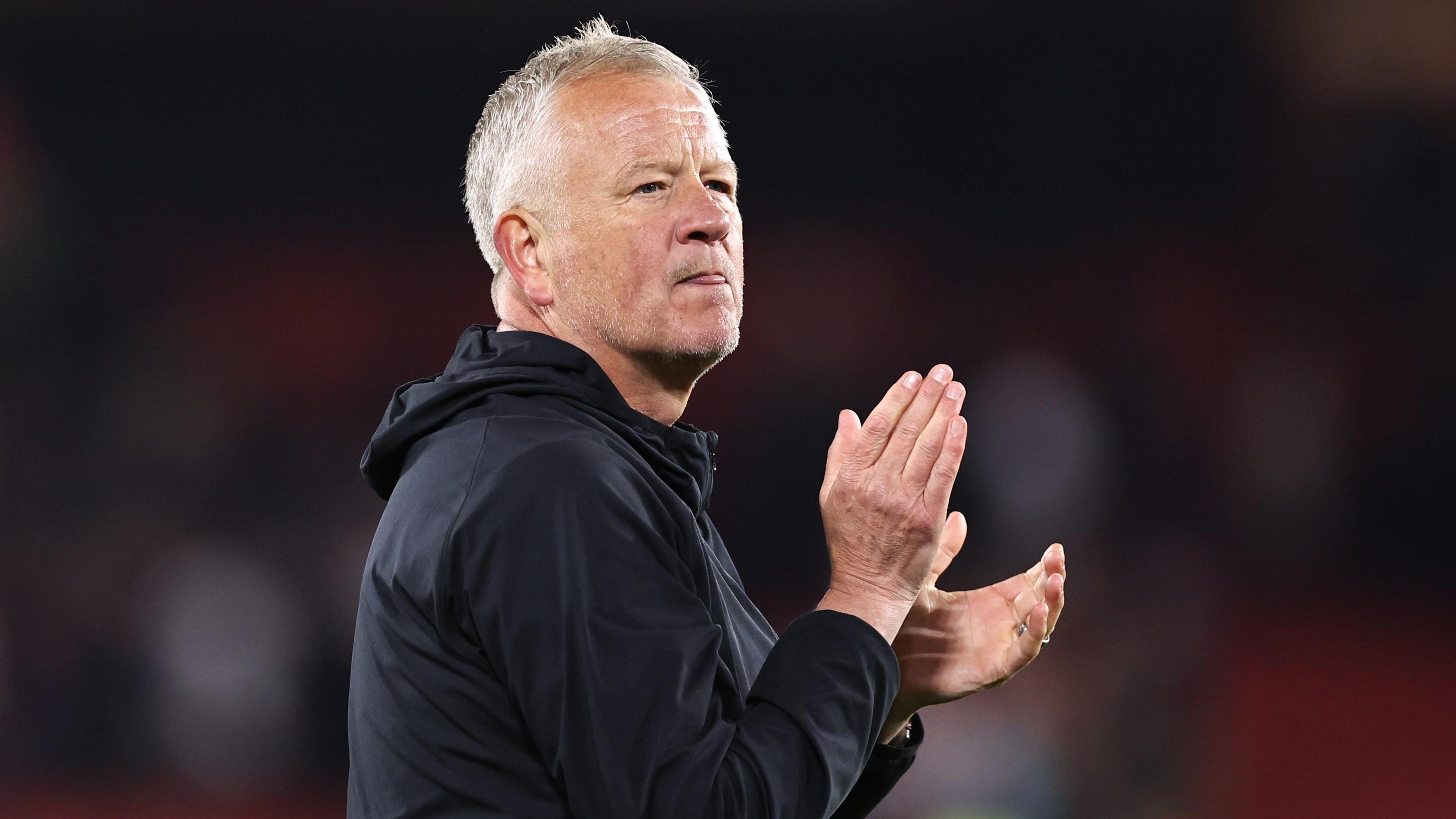 Chris Wilder, a man in his fifties, wearing a dark top. He is looking into the far distance and applauding.