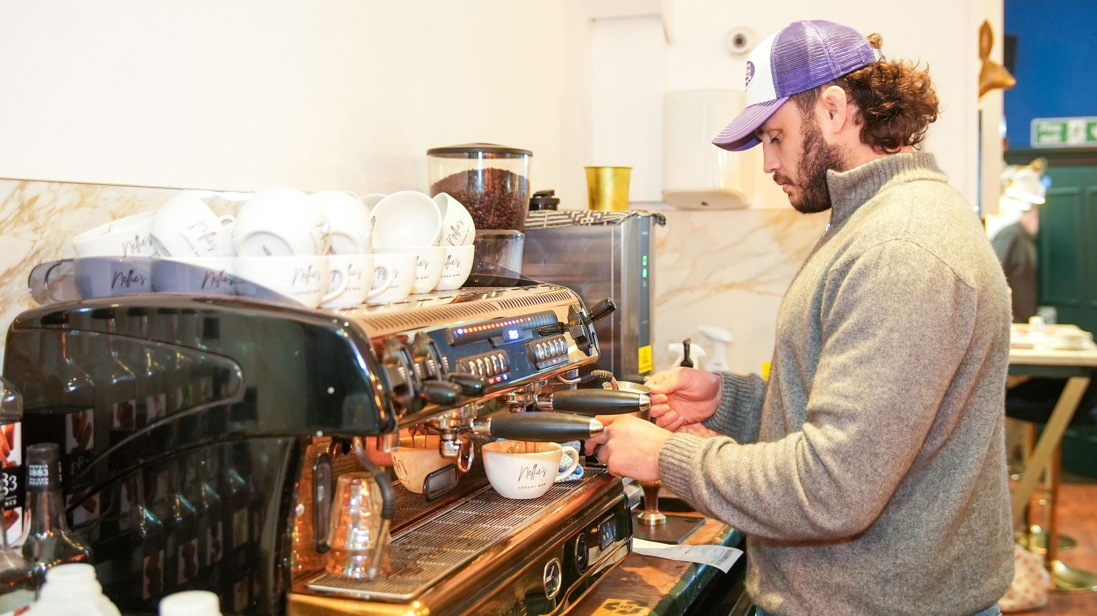 A man concentrating making coffees using a coffee machine. He's wear a grey jumper with a purple hat. The coffee machine has lots of cups above it with coffee beans stored alongside. 