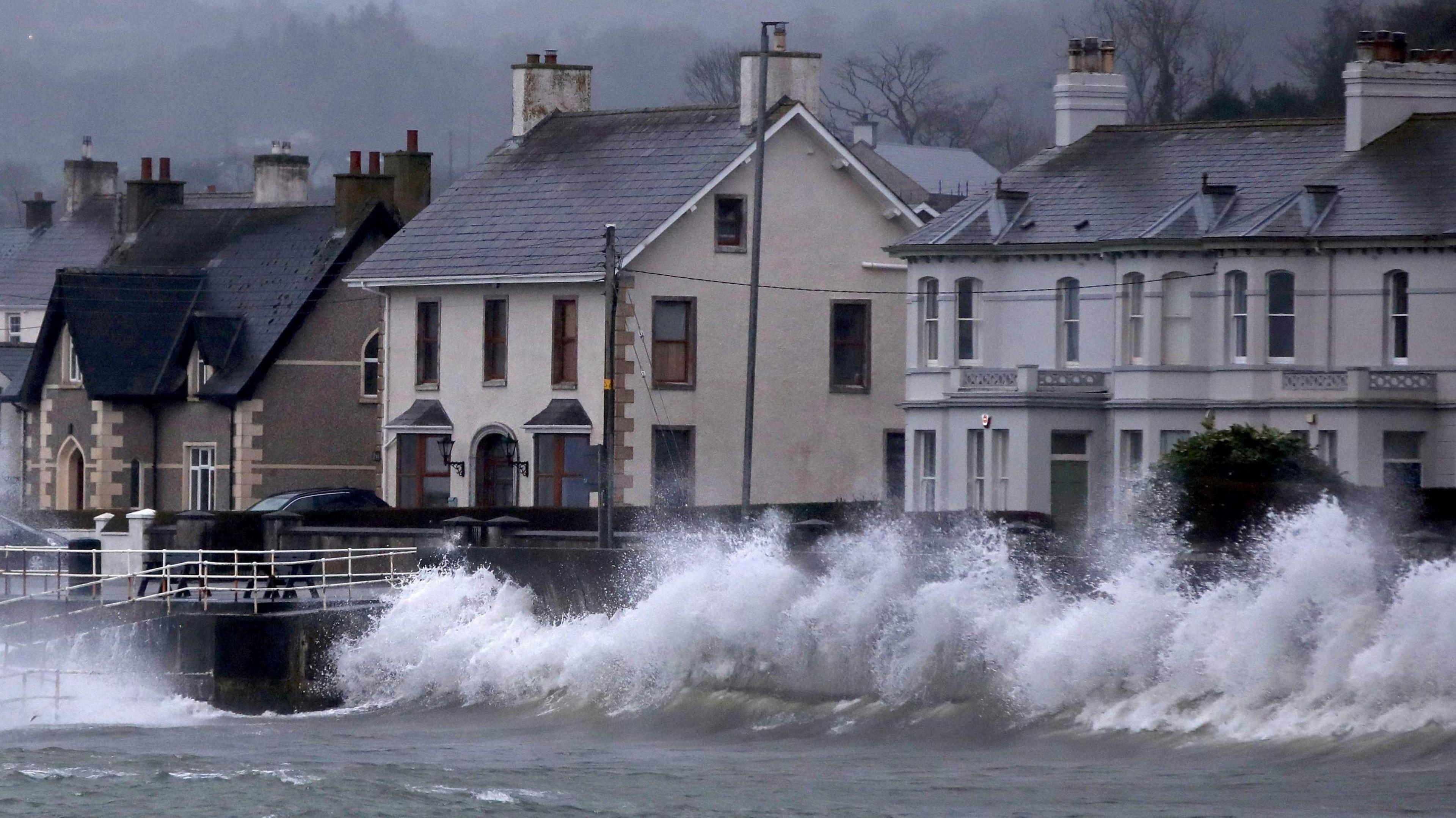 A row of houses by the sea. There are large waves hitting the wall between the sea and the houses.