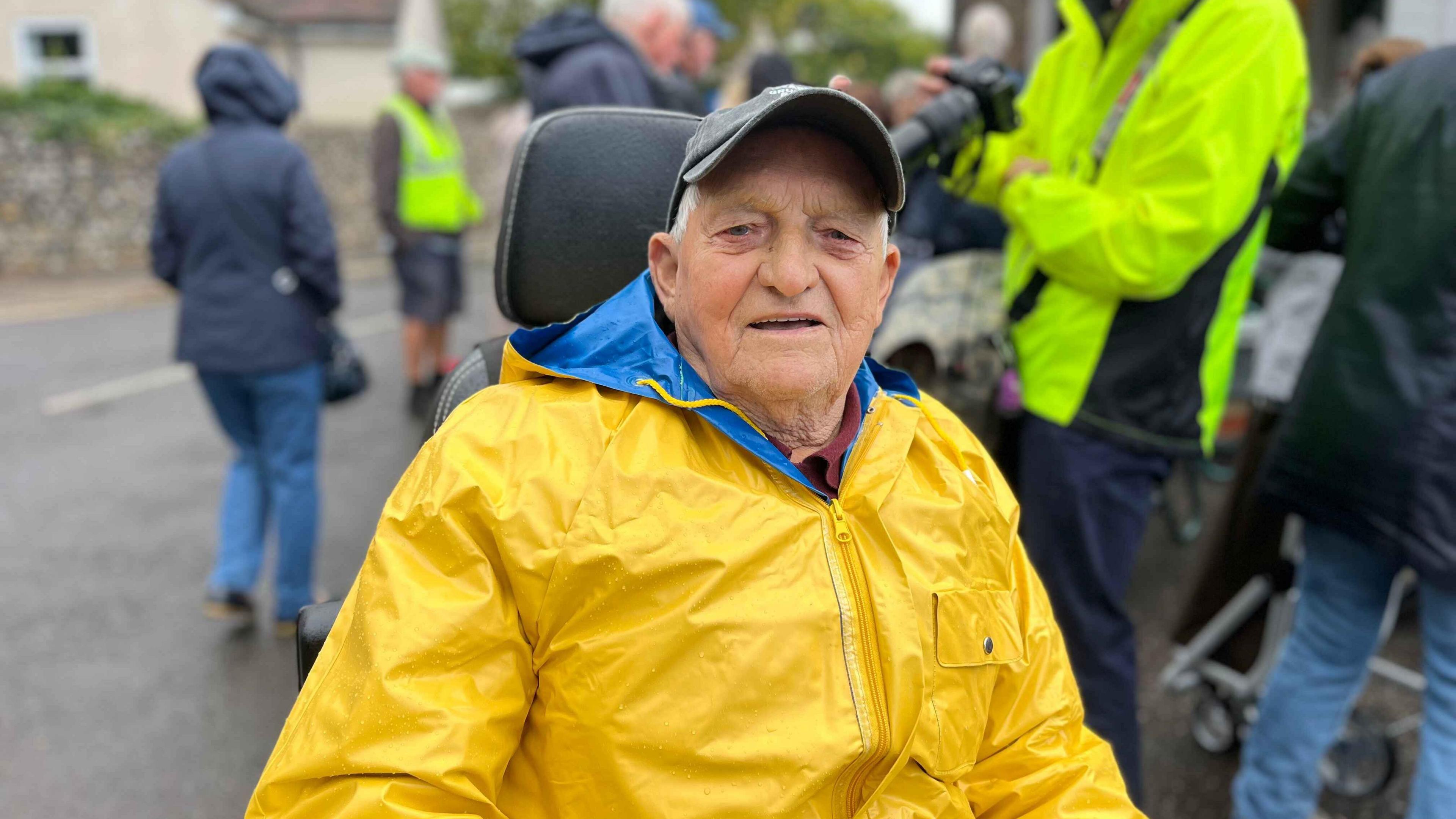 John Ball sitting in his mobility scooter smiling at the camera wearing a yellow raincoat and dark cap
