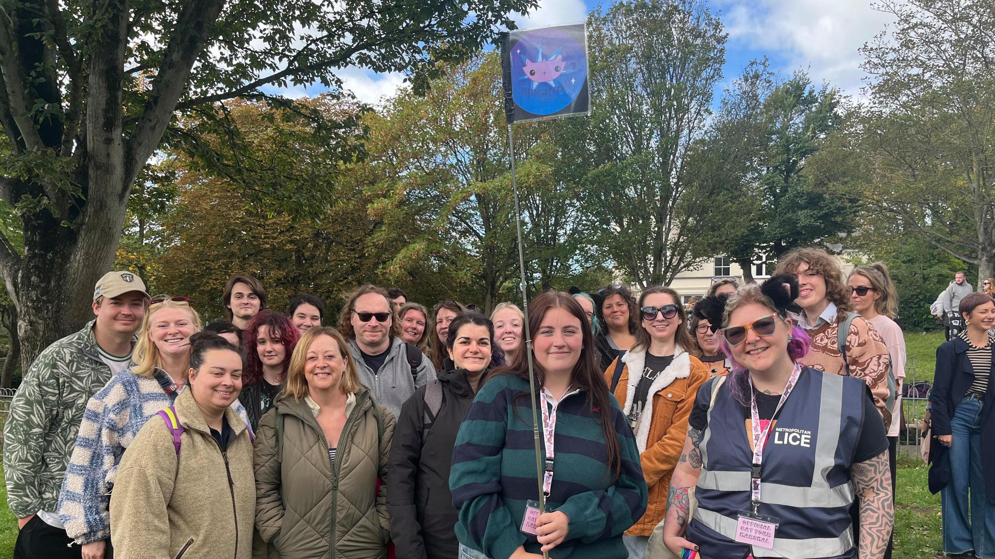 A crowd of over 20 people pose for a photo alongside volunteer marshal Nia Wildblood in an open green space next to a tree. One woman (centred) holds a flagpole with a small, square flag on top showing the Brighton Cat Tour logo. People smile for the photo while looking directly at the lens.