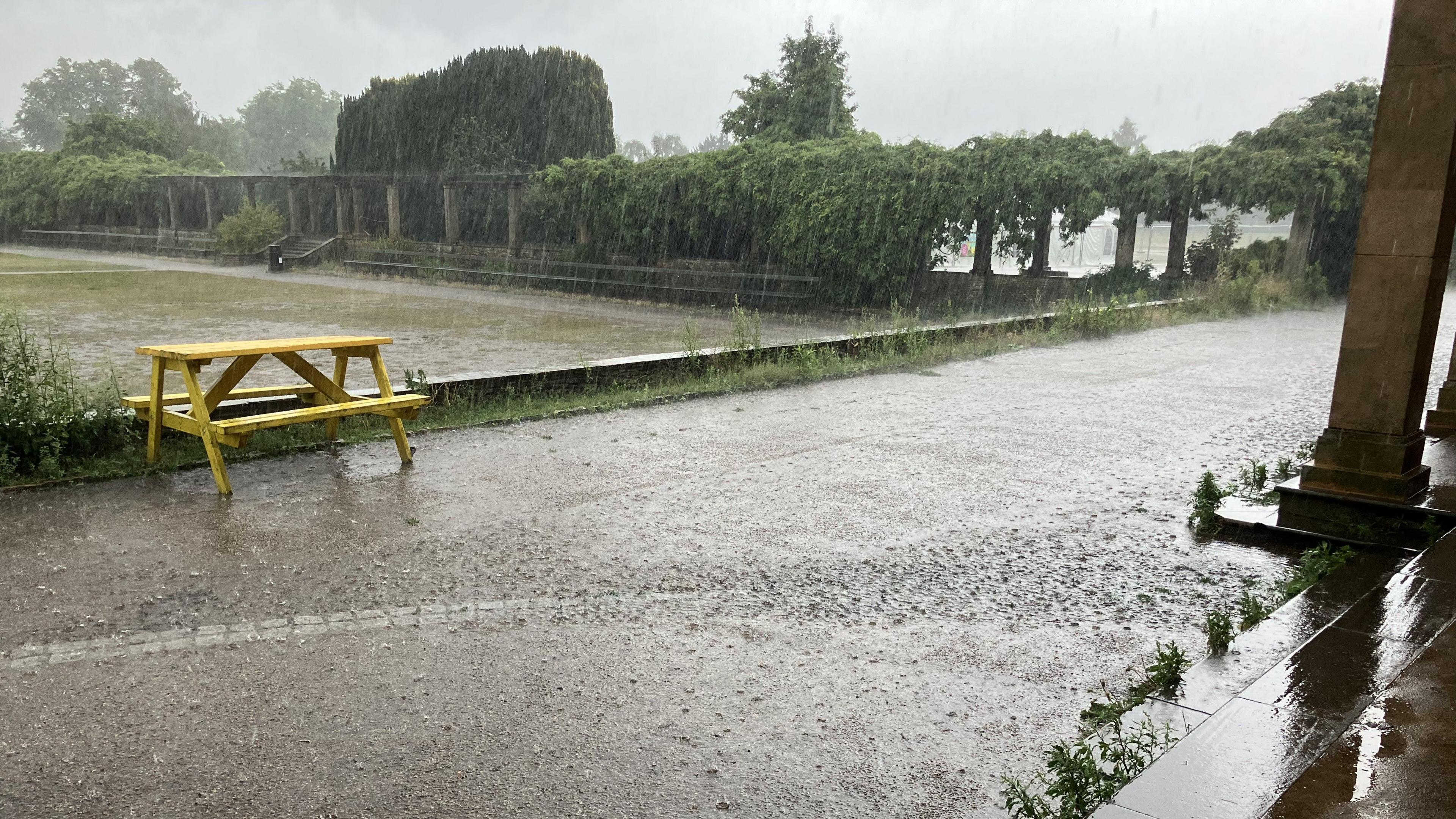 A heavy downpour of rain hits a park. An area of concrete can be seen covered in rainwater.
