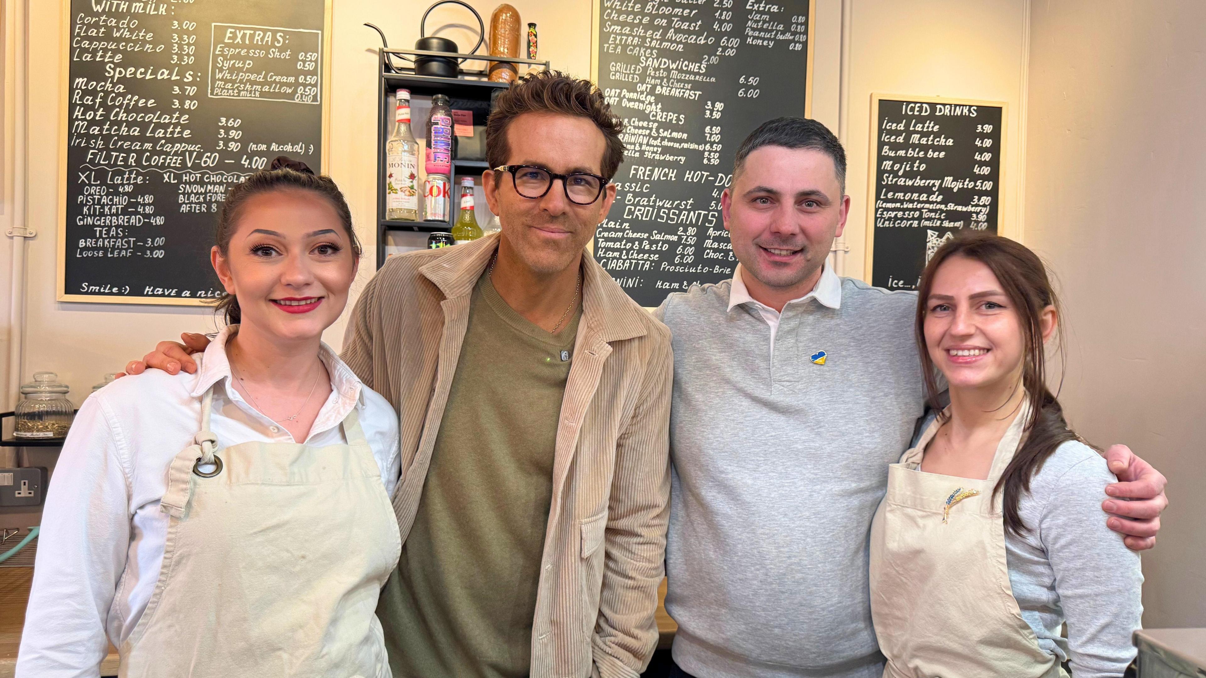 Four people in a cafe. From left to right is a young woman, Ryan Reyonolds, Yaroslav Izviekov and another young woman. Three blackboards with the cafe menu can be seen in the background, and the two women are wearing cream aprons. 