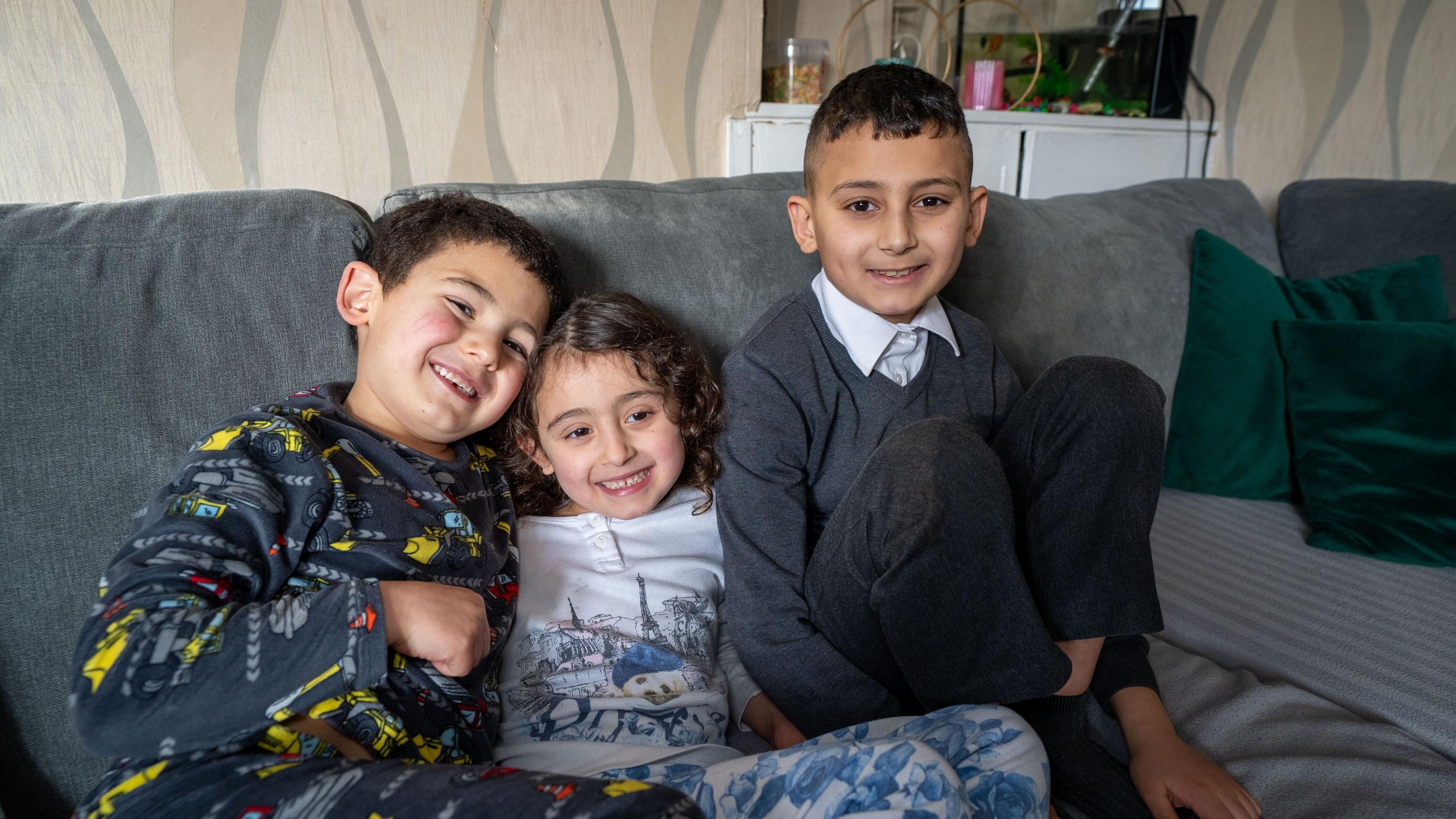 Three young smiling children sit on a couch in a living room. They are bunched up close together. A girl in the middle and a boy on her right are wearing pyjamas, while an older boy on her left is wearing his school uniform. They all have dark hair