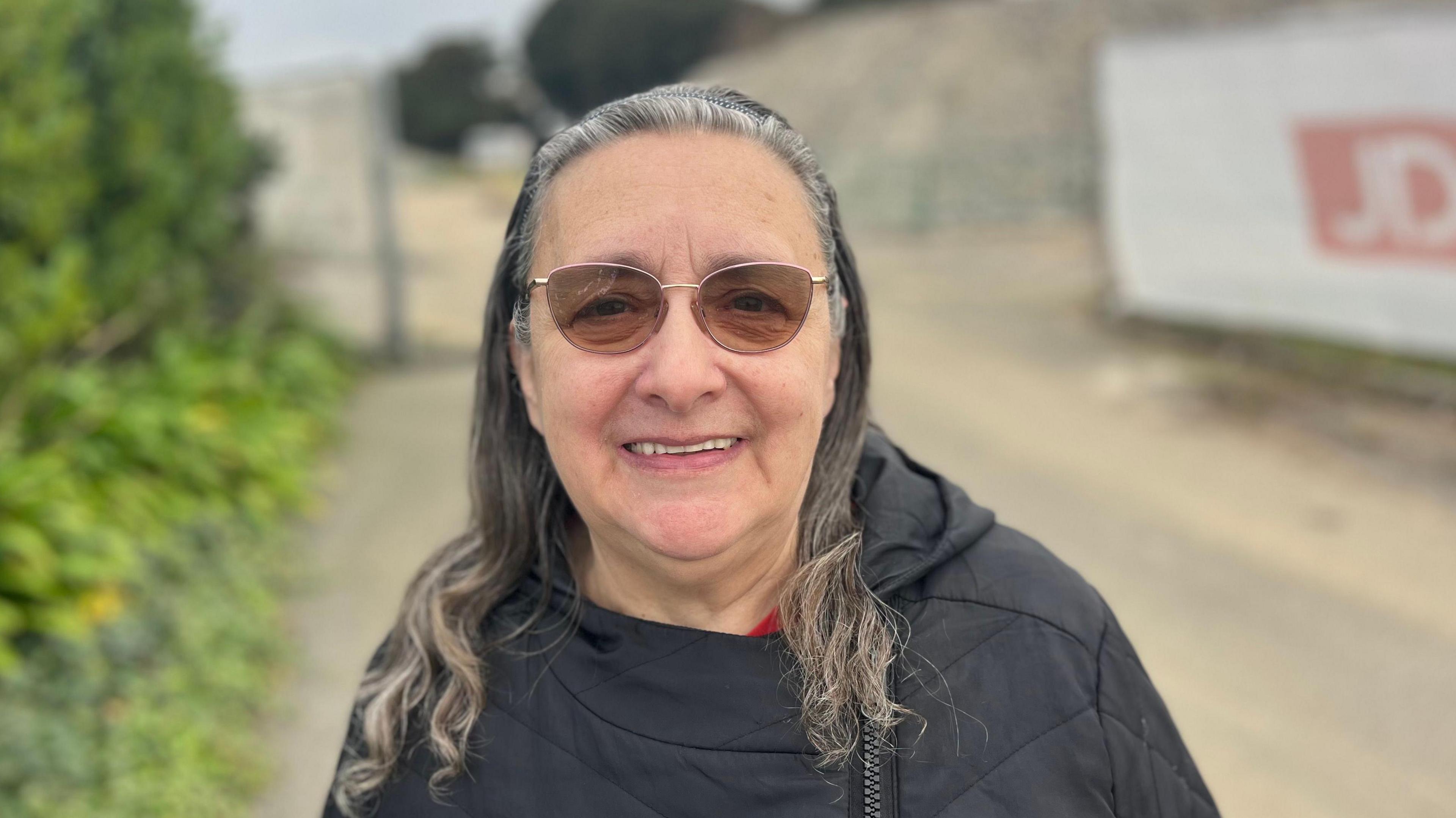 Jill is an older woman with long grey hair. She is smiling at the camera. She is wearing a black coat and brown glasses. She is standing in front of an out-of-focus construction site, with a green bush to the left of the photo and a white fence to the right.