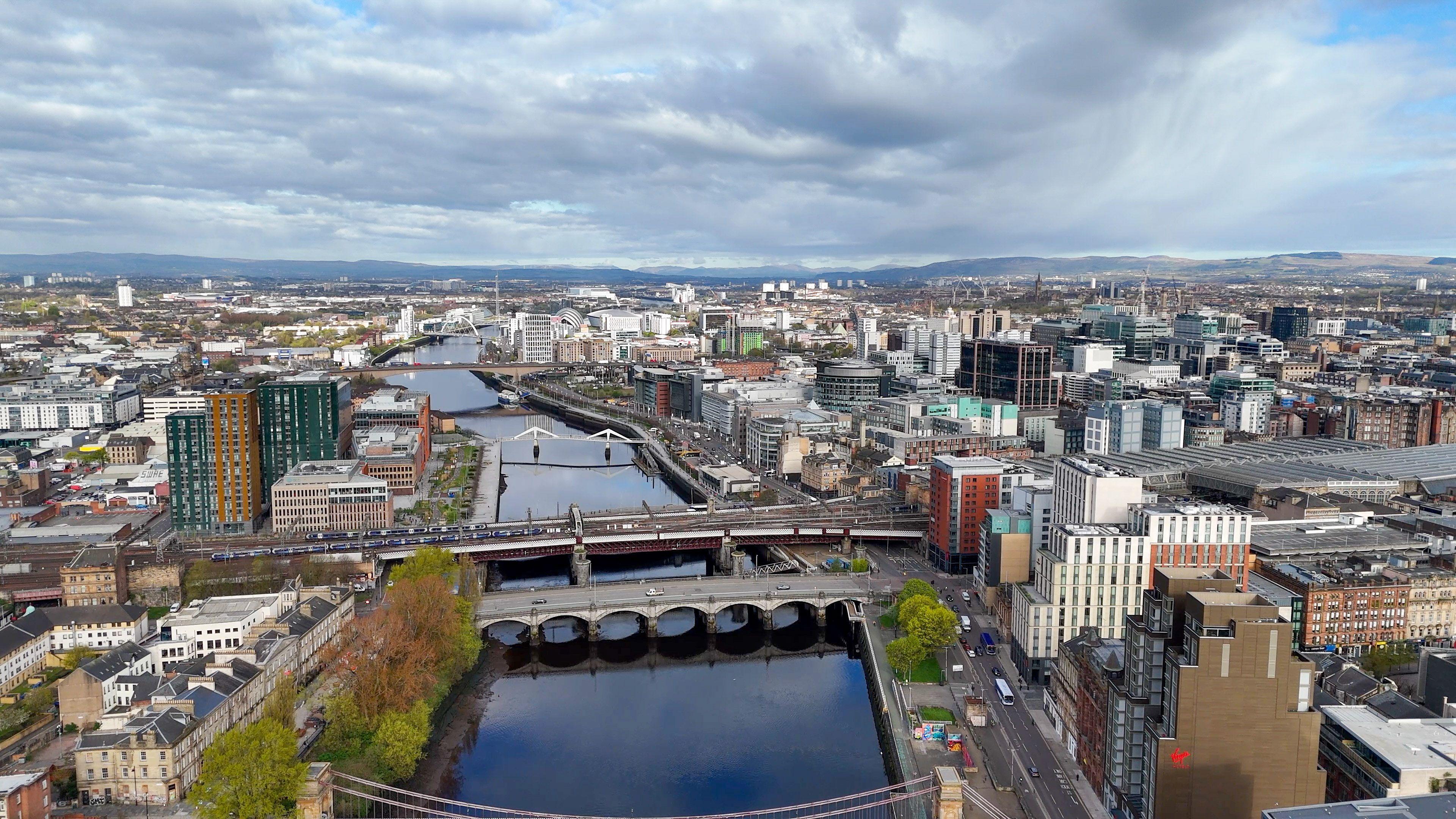 Aerial view of Glasgow city centre showing the River Clyde, multiple bridges, railway lines and modern office buildings beneath dramatic clouds stretching across the skyline.