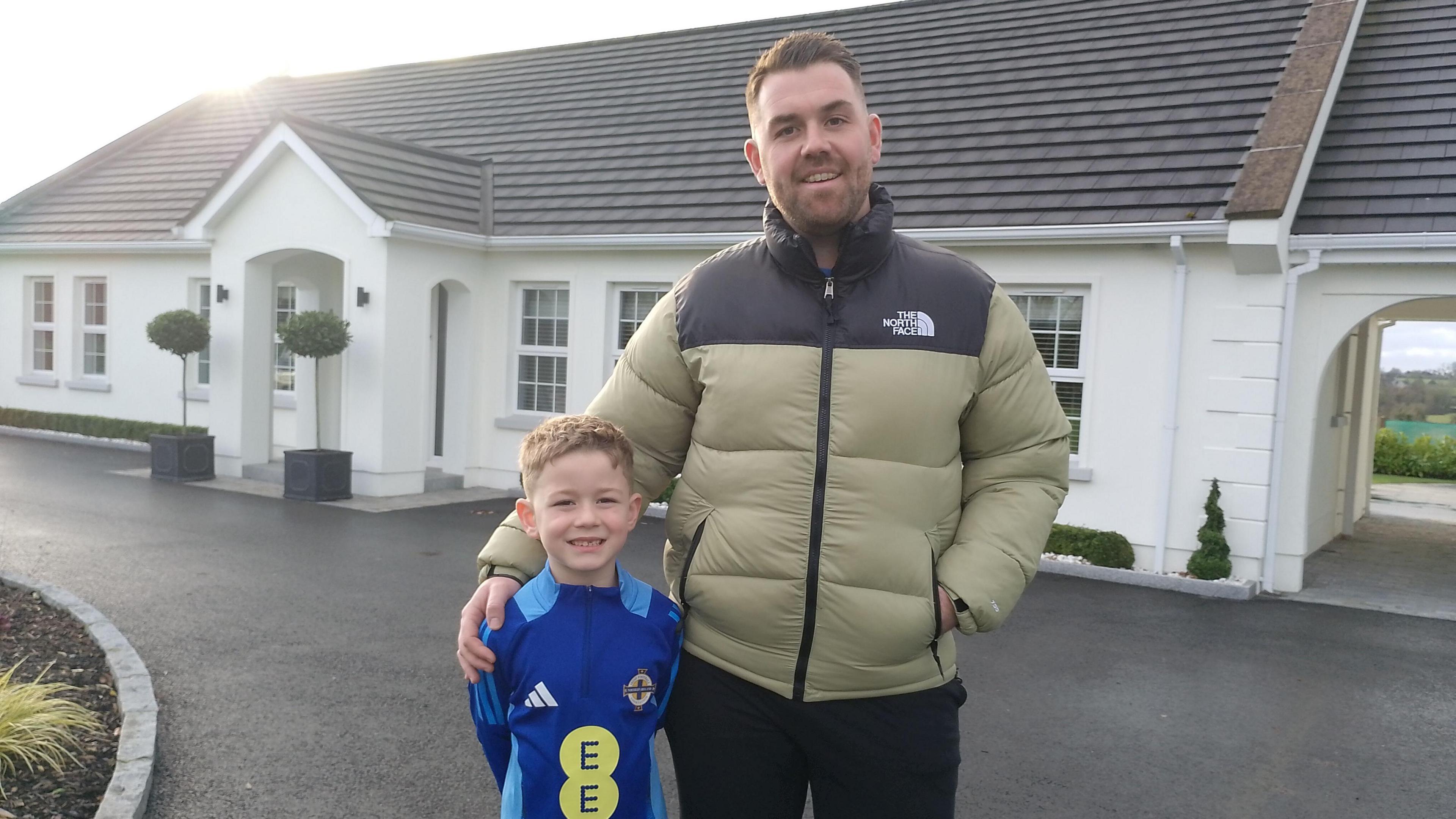 A man and his son stood outside their white home. The father is wearing a khaki winter coat and the young boy under his arm is wearing a blue Northern Ireland football kit. Both are smiling at the camera