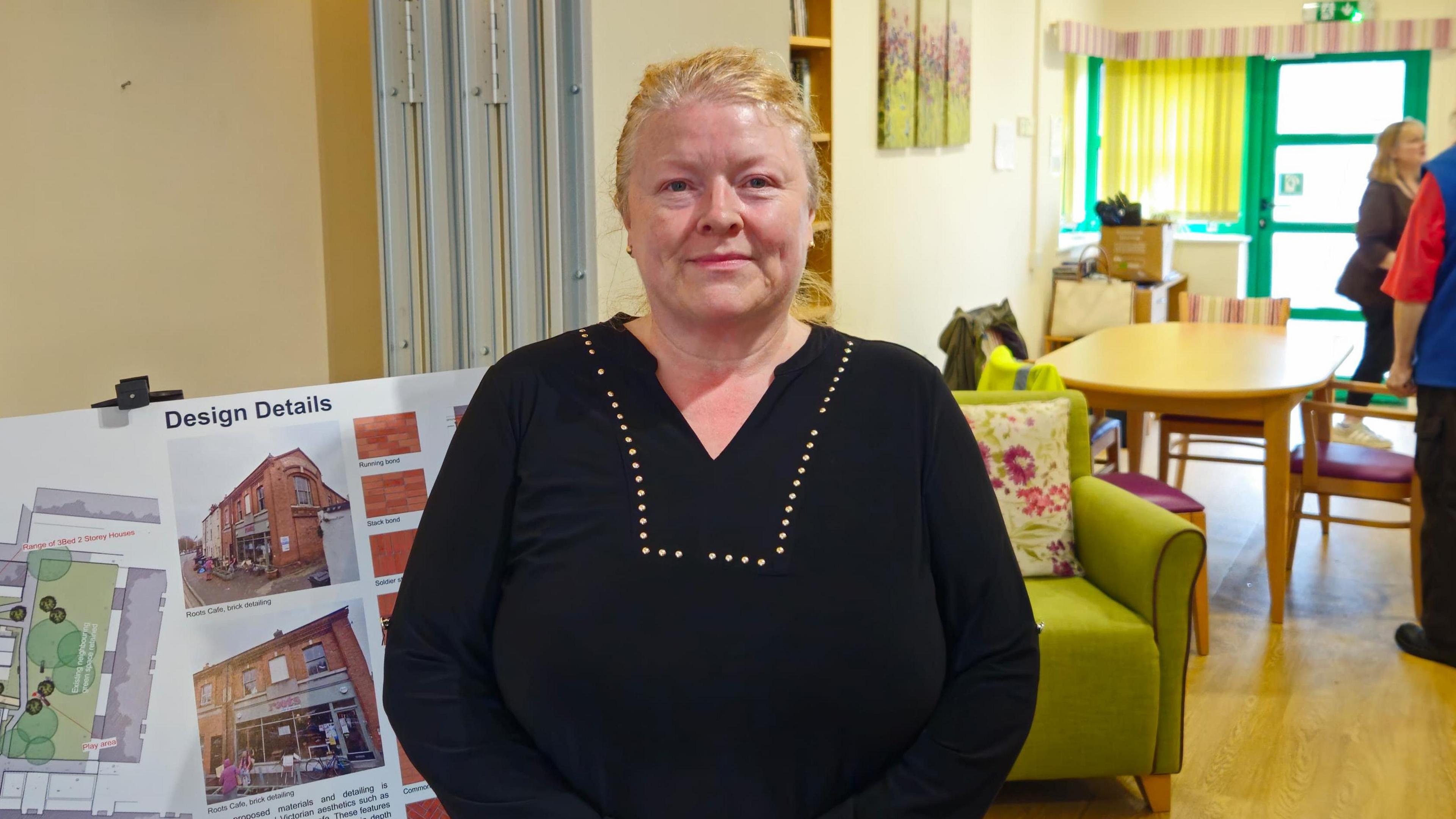 A photo of a white woman with blond hair, She is wearing a black top with beads around the neck area, She is stood in front of a board with plans on and a green chair