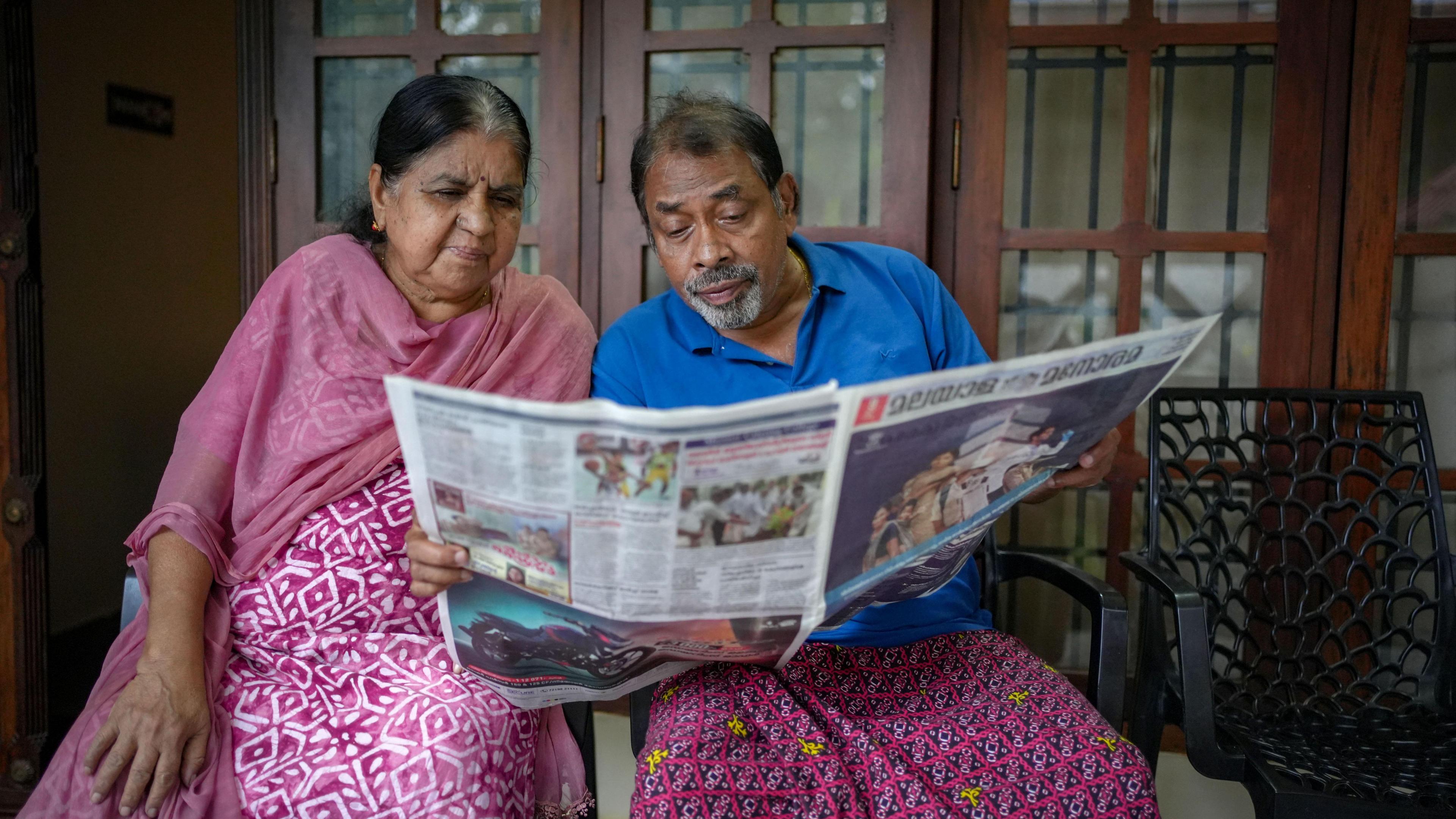 TO Dominic and his wife sit side by side indoors, holding and reading a newspaper together.